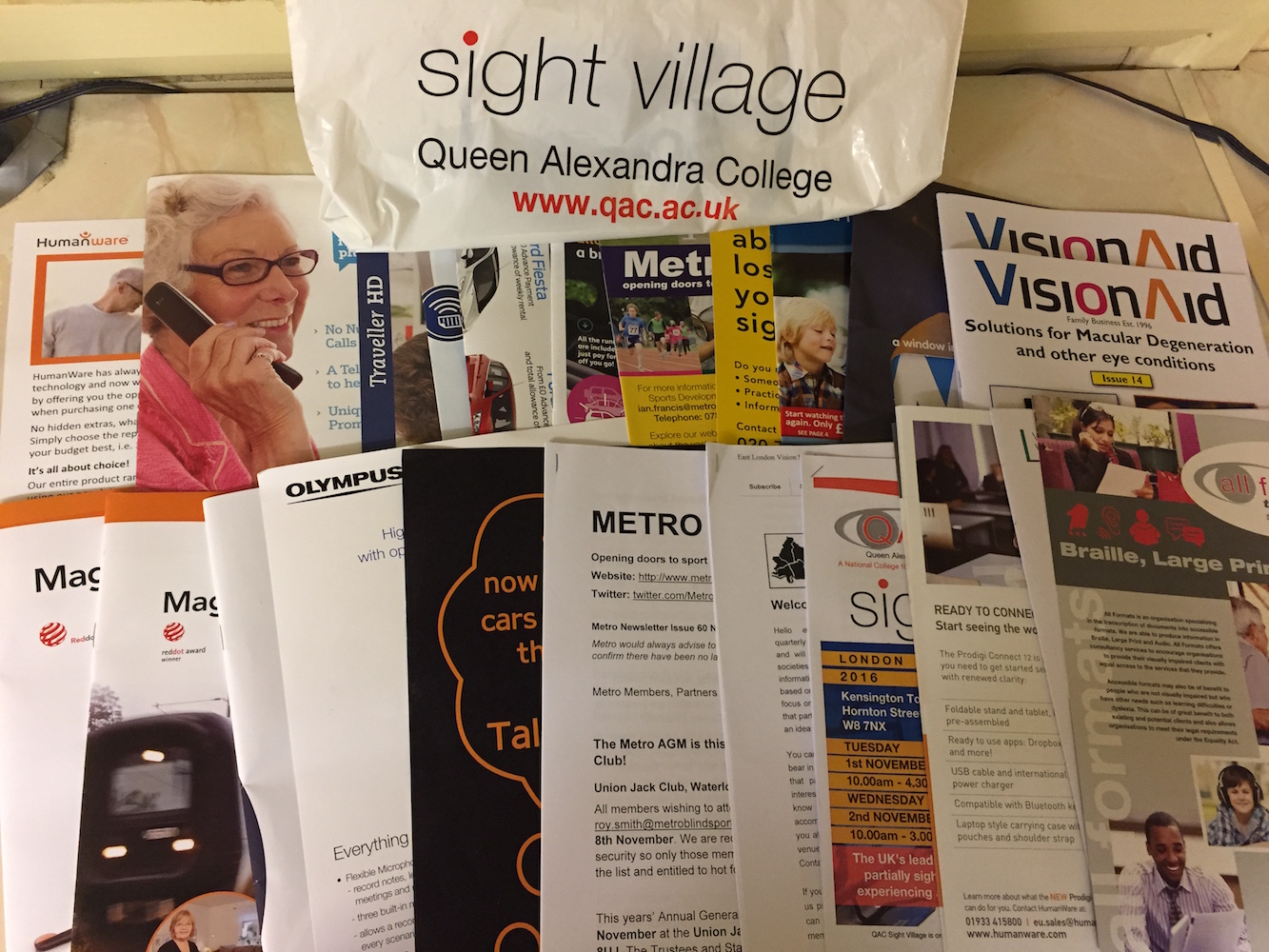Large collection of booklets and leaflets from Sight Village for various organisations. At the back is a bag that says Sight Village, Queen Alexandra College, www.qac.ac.uk.