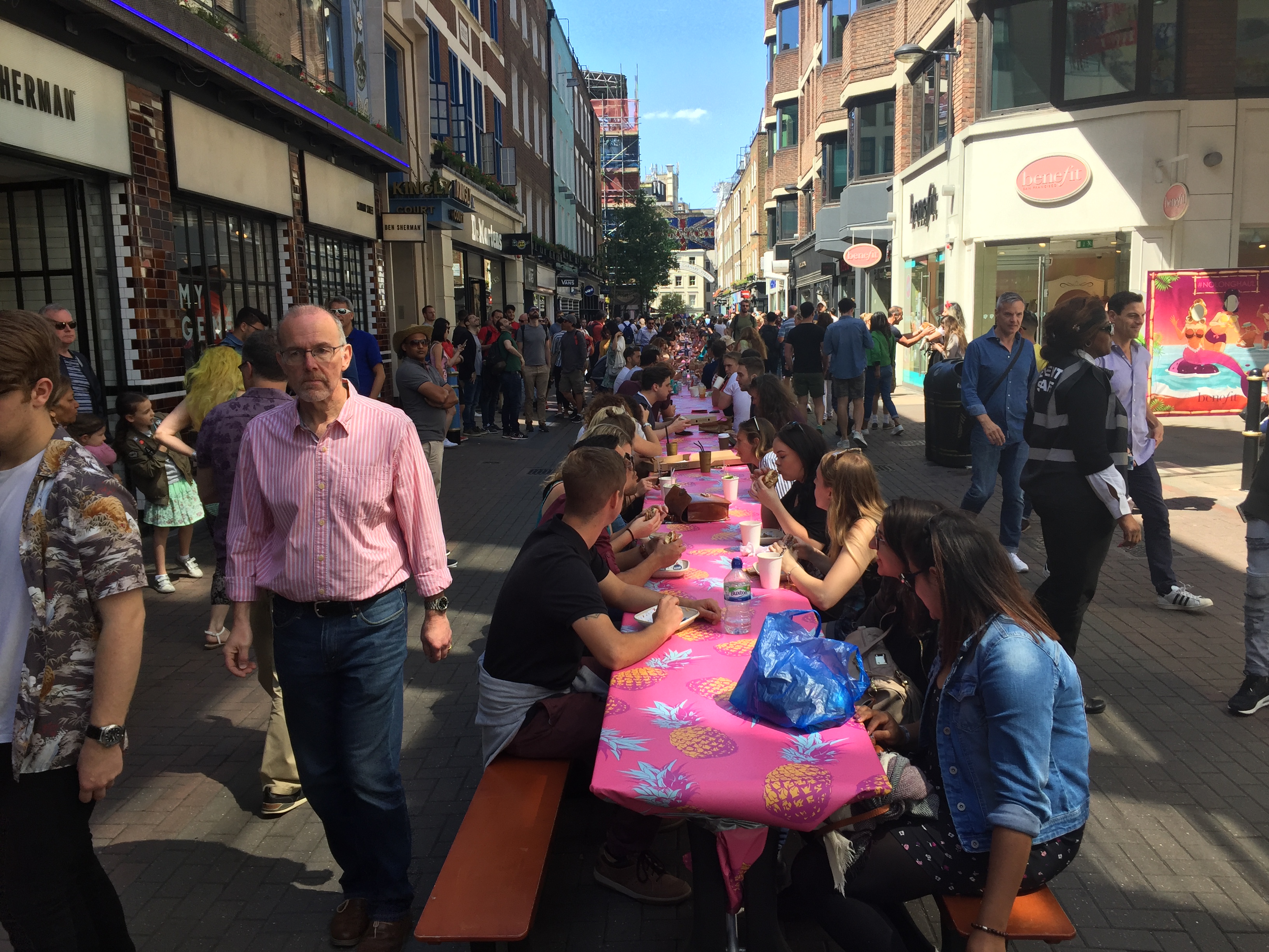 A long, narrow table stretches down the narrow Carnaby shopping street, with people sat on each side enjoying their picnics. The tablecloth is pink with images of pineapples. Many more people are walking down the street on each side.