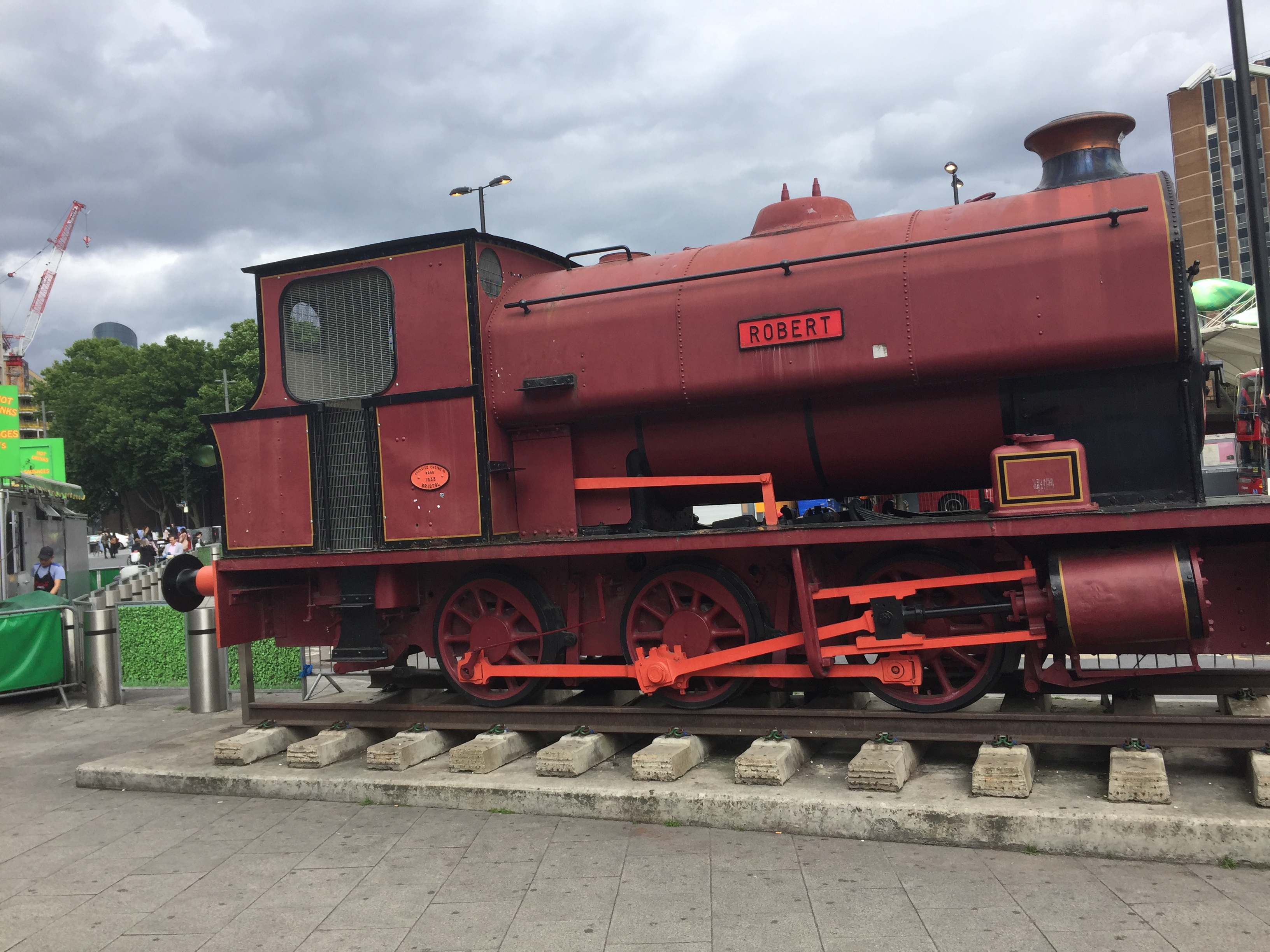 A large red steam train, with the name Robert in a small plaque on the side, sits on a small section of railway track placed outside Stratford Station.