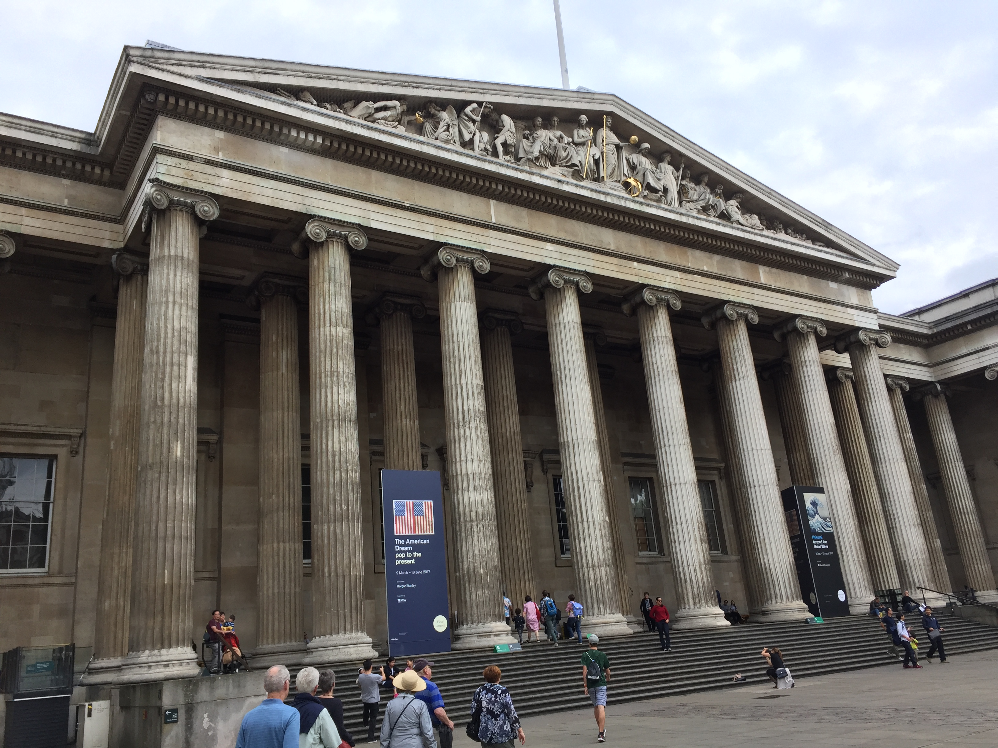 The entrance to the British Museum. At the top of the wide steps are 8 tall pillars, spaced along the width of the steps, above which is a triangular roof section containing various carved figures.