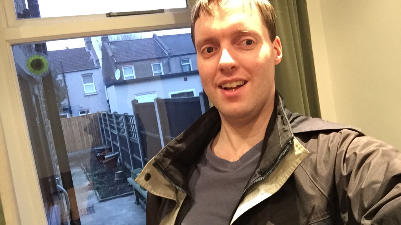 Selfie photo of me wearing a coat, standing in front of a window looking out at the paved back garden, with the backs of other houses beyond the wooden fencing.
