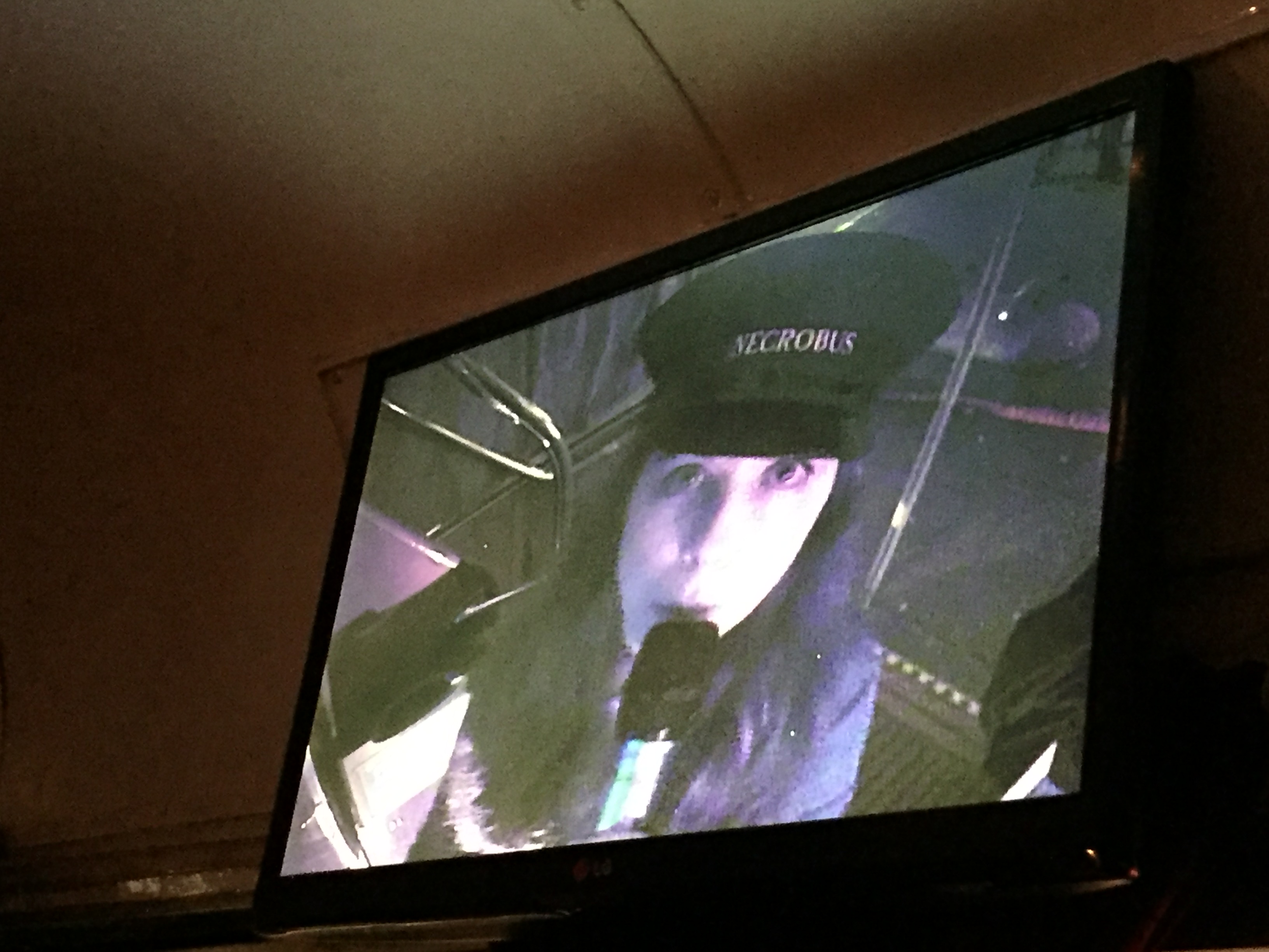 Lady tour guide on the bus, wearing a black hat with the word Necrobus on it and dark clothes. She is holding a microphone and looking at the camera, and we are viewing her on a monitor on the upper deck of the bus.
