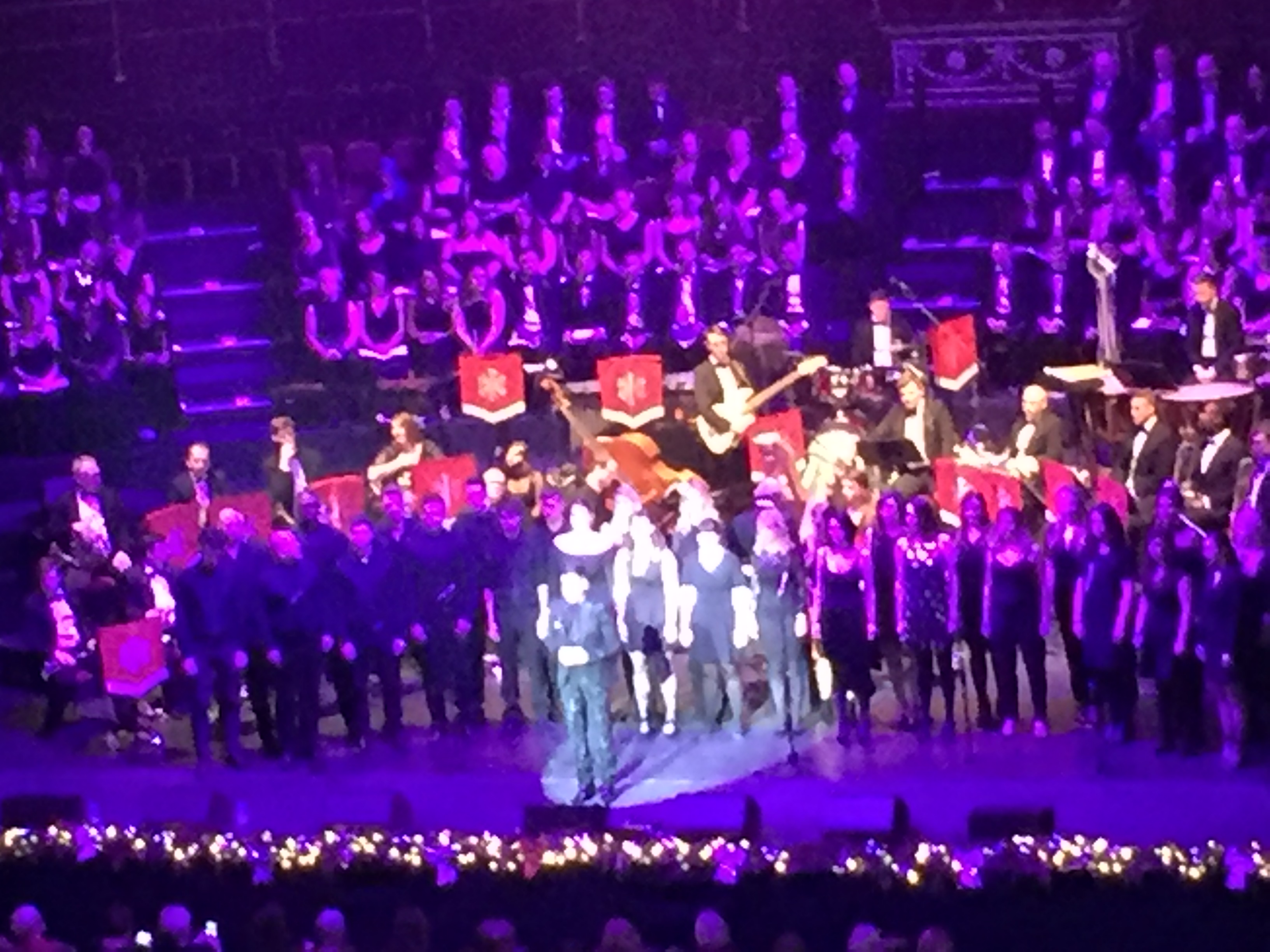 Performers on the stage. From front to back there is a solo singer, a line of backing singers, the band in a semi-circle, and a choir spanning multiple rows curving around the back of the stage.