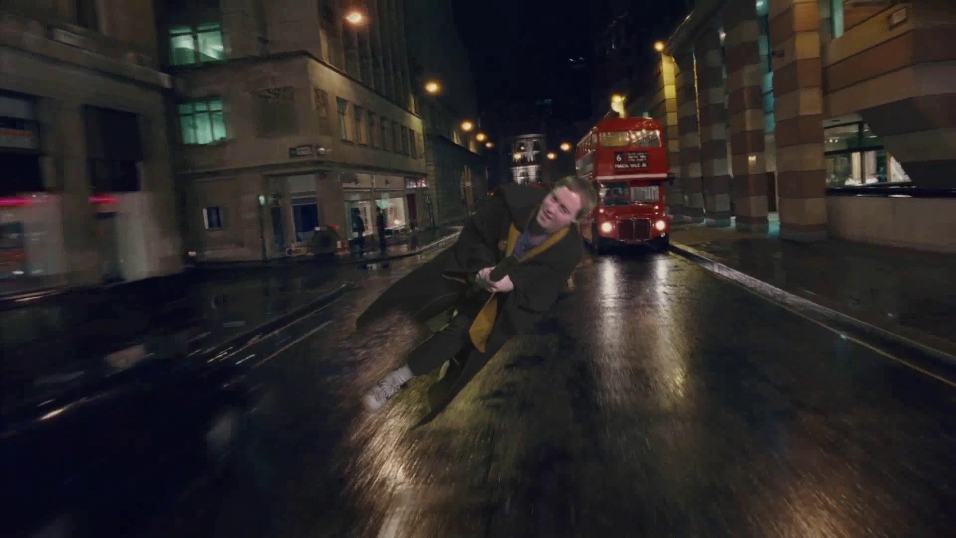 Glen in a brown robe flying on a broomstick, down the middle of a busy London road at night, with a red London bus in the background.