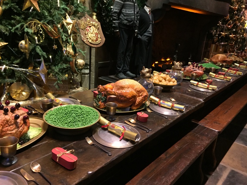 In front of a large Christmas tree is a long wooden table laid out for Christmas dinner, with large cooked birds, bowls of roast potatoes and peas, while each diner gets a golden cracker, gold drinking goblet, gold cutlery, a silver plate, and a small red present tied with gold ribbon.