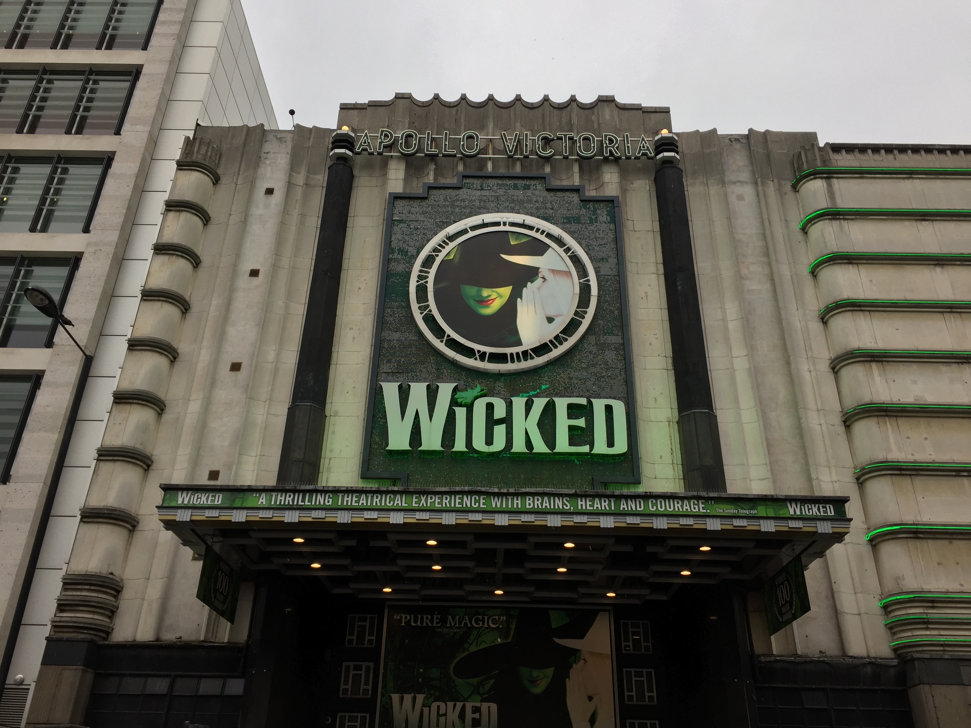 Wicked sign above the Apollo Victoria Theatre, with Elphaba, the green coloured Wicked Witch of the West, in the middle of a clock face, above the show's title in large letters. Along the front of the narrow canopy above the theatre entrance is white text on a green background, which reads: "Wicked - A thrilling theatrical experience with brains, heart and courage."