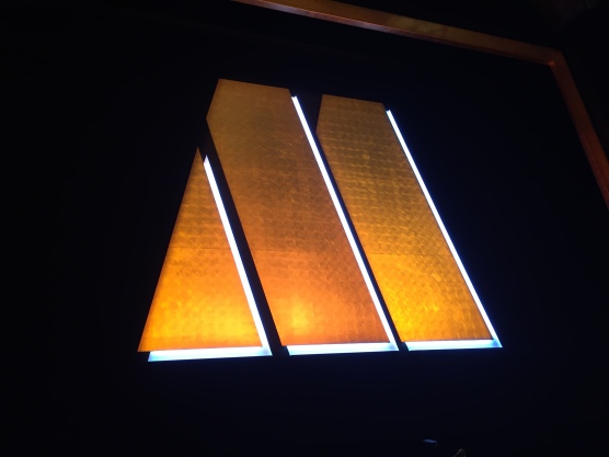 The large golden letter M logo for Motown, made up of a triangle then 2 thick diagonal bars, lit up on the black stage curtain.