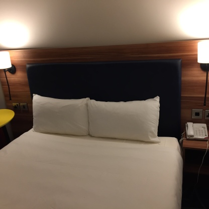 Double bed in my hotel room, with a lamp on the wall on each side, and a telephone on the bedside table.