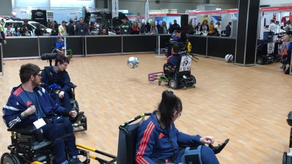 Powerchair Football at Naidex. The ball has been launched into the air as it travels towards the goal.