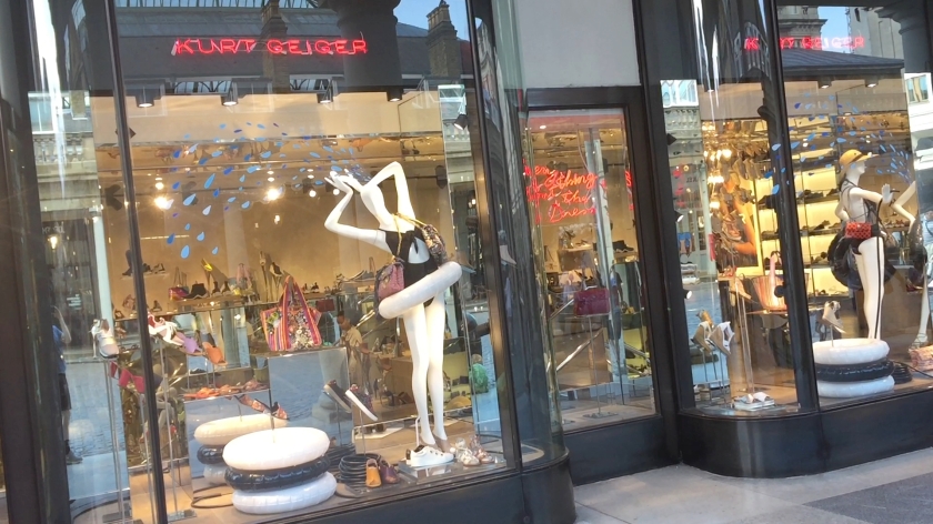 Shop window in Covent Garden with a variety of clothing and accessories. The most striking object is a bright white mannequin wearing a black bikini and a white rubber ring around the waist. The shop name above the window and on the door way is in curly text lit up in an orangey-red colour.
