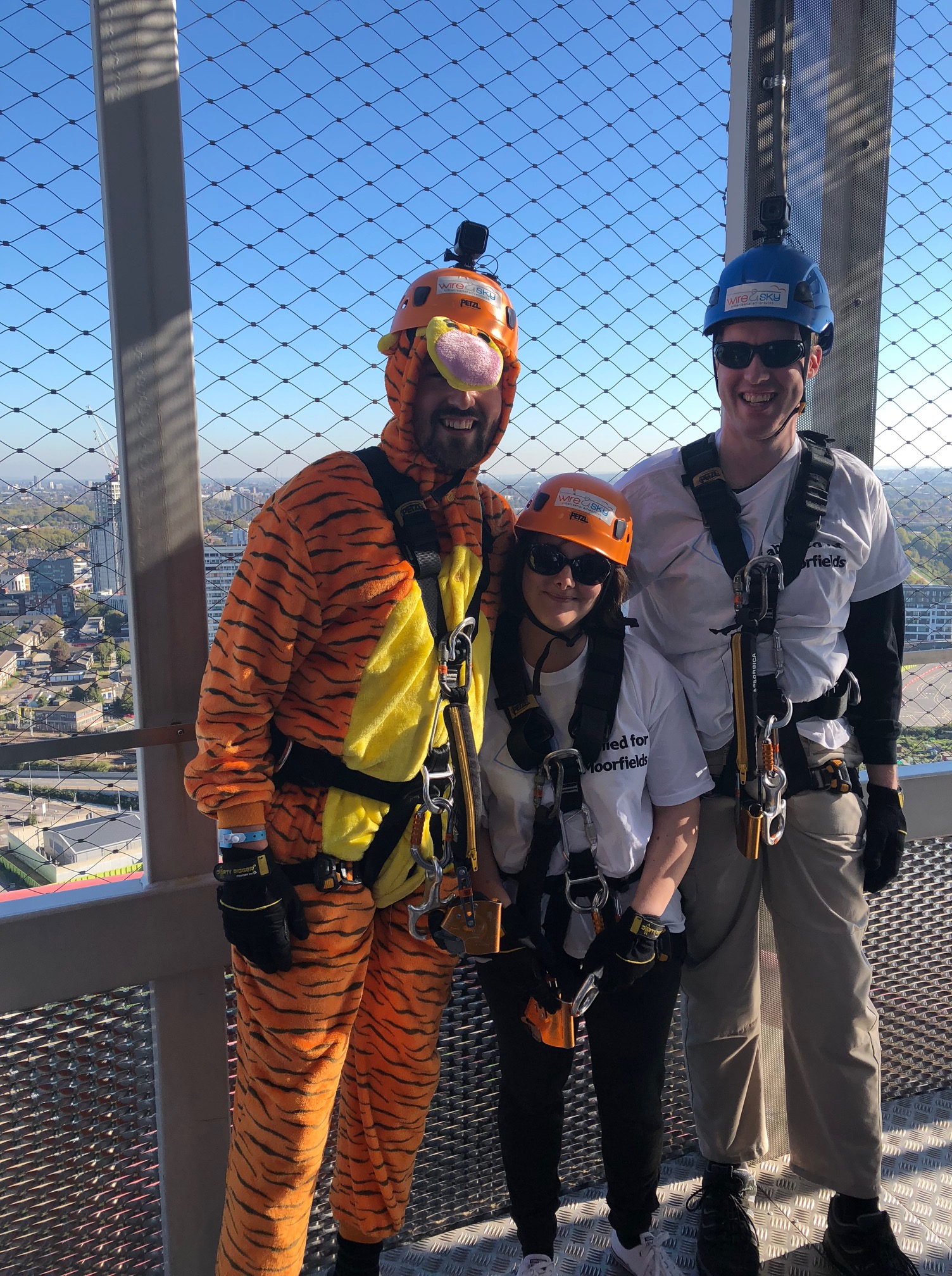 Matt, Claire and Glen ready to abseil in their harnesses and helmets. Matt and Glen also have GoPro cameras attached to their helmets. Glen and Claire are wearing their Moorfields Eye Charity t-shirts and normal trousers, but Matt is wearing an orange furry Tigger bodysuit, to look like the character from the Winnie The Pooh stories. It has black tiger stripes, a yellow chest, and a big round grey nose poking out from under his helmet.