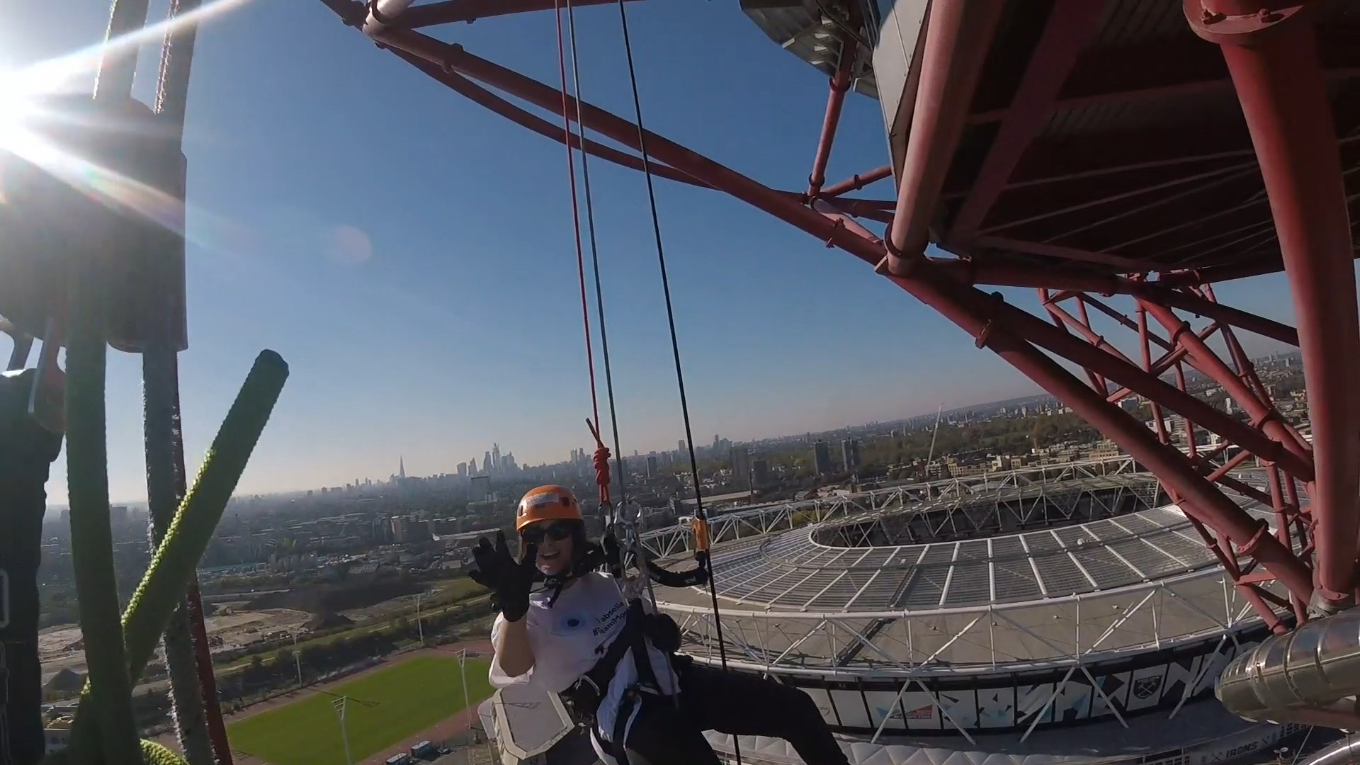 Headcam view to the right of the tower, showing fellow abseiler Claire smiling and waving, with the large round Olympic Stadium behind her, and the London skyline stretching into the distance.