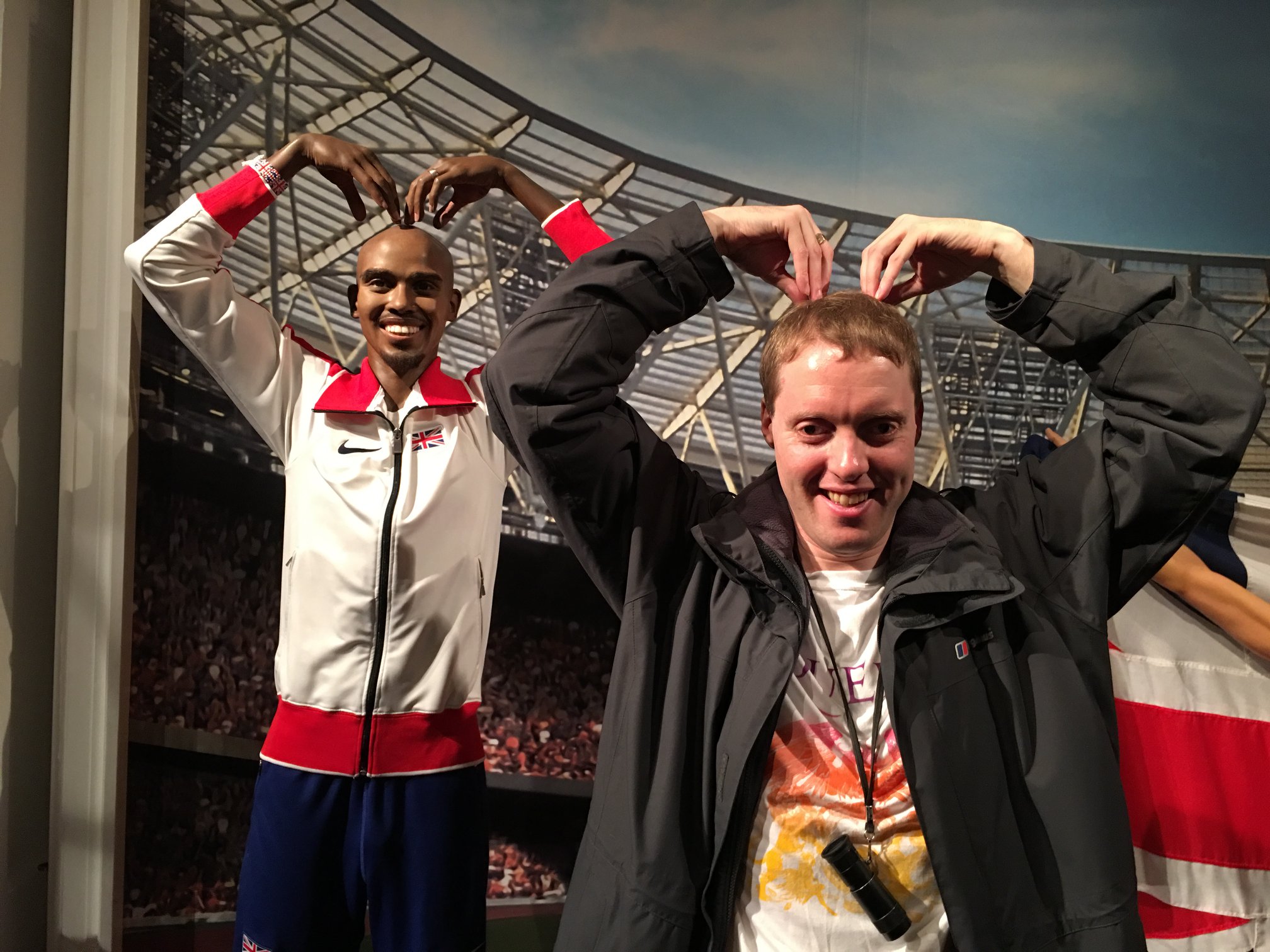 Glen next to a waxwork of Mo Farah, copying the Mobot stance that Mo is doing, placing both hands on top of his head with his fingers pointing downwards.