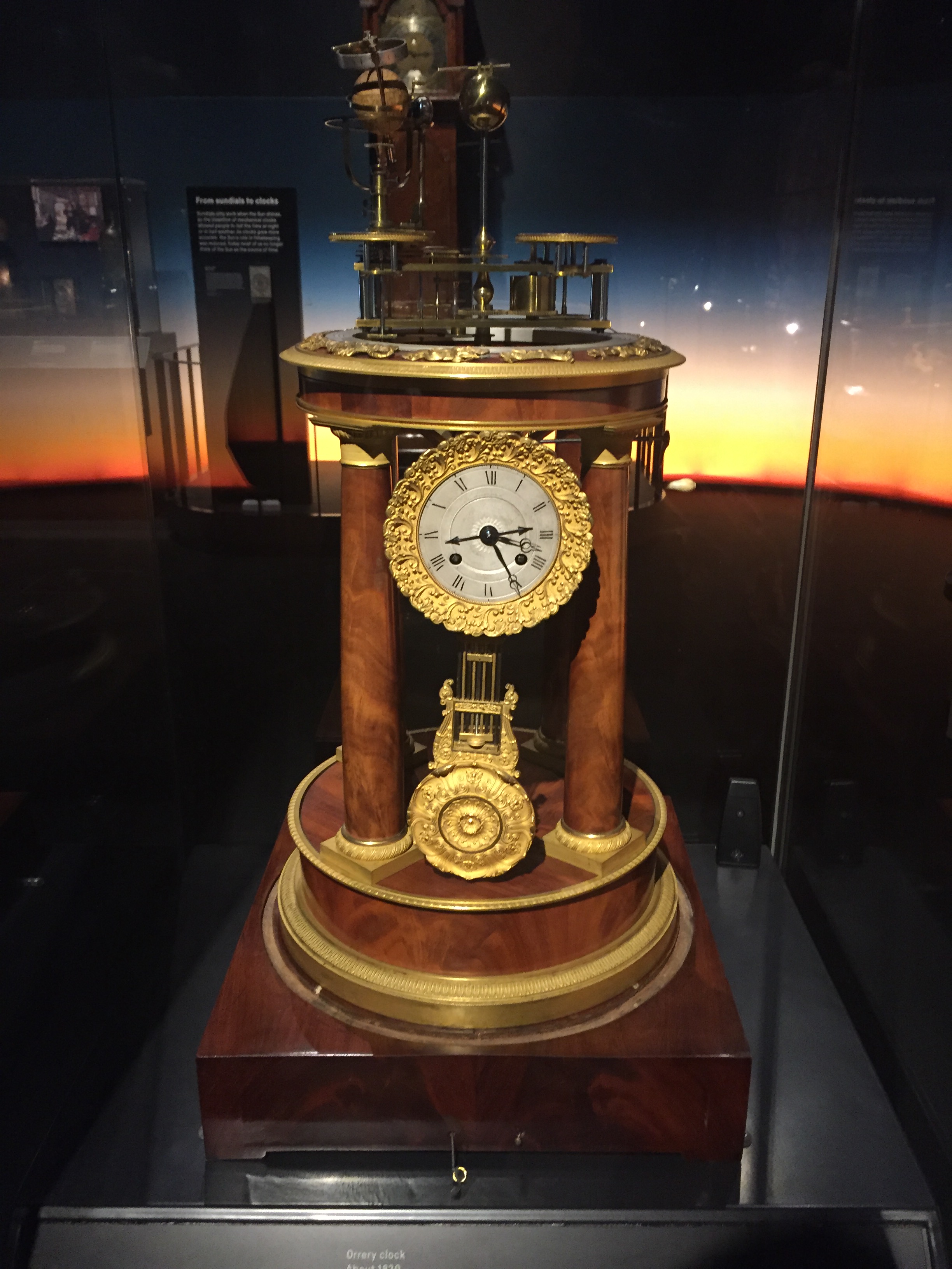 An ornate desk clock with a large round base, a detailed gold border around the face. and a mechanism on the round top that can be used to determine the time using the sun's position.