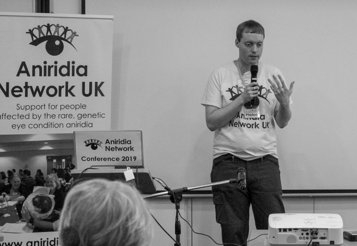 A black and white photo of Glen standing and talking into a microphone. He is wearing a white t-shirt with the Aniridia Network logo on it, while next to him is a tall free-standing banner for Aniridia Network UK, and a laptop on a podium with an Aniridia Network Conference 2019 sign on its open lid.