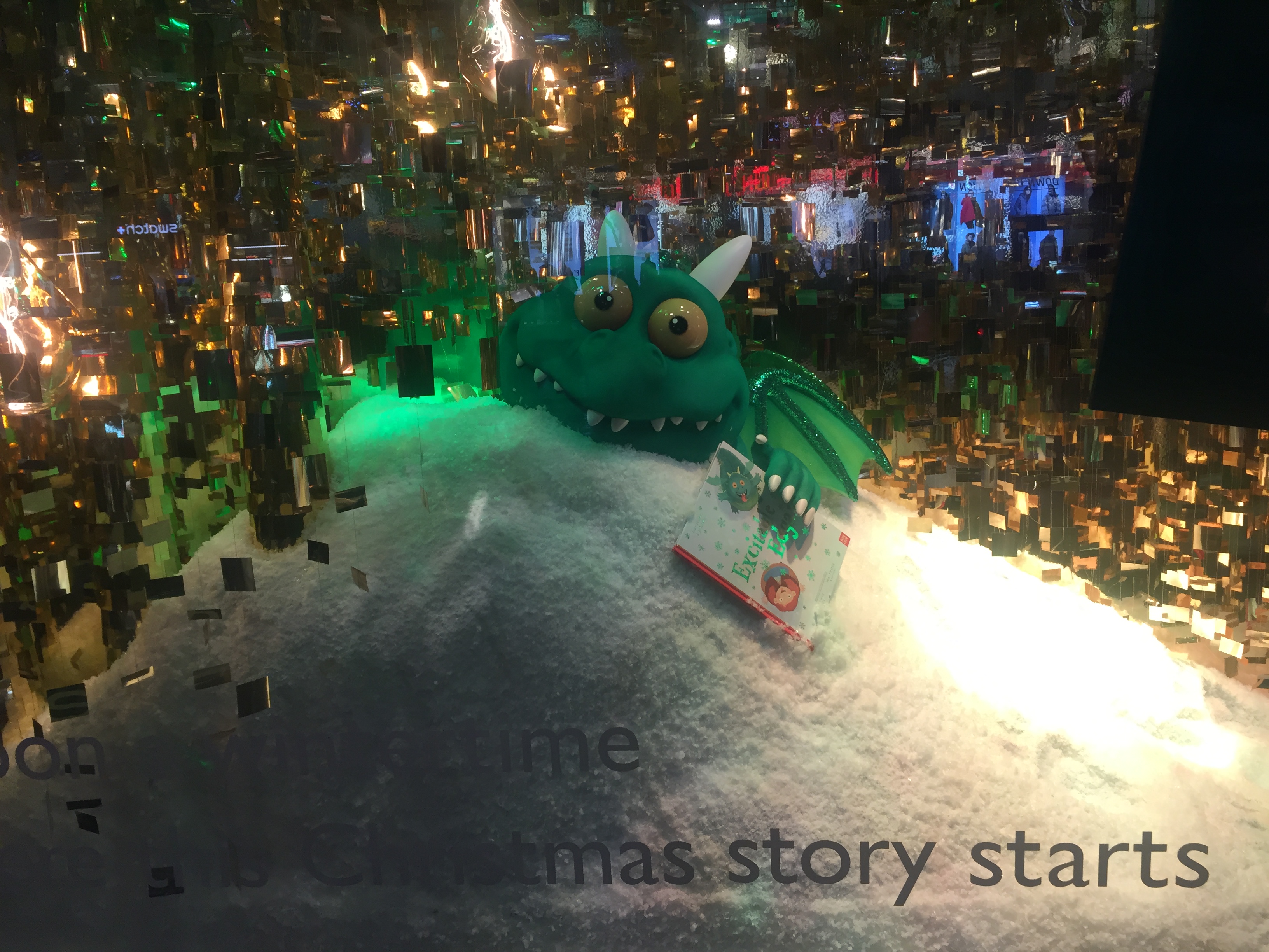 John Lewis window display showing Edgar The Dragon buried in a pile of snow with only his head showing. He has green skin, white horns and claws, yellow eyes with black pupils, and sharp white teeth. He is holding a book called Excitable Edgar in one hand, with an image of him on the cover.