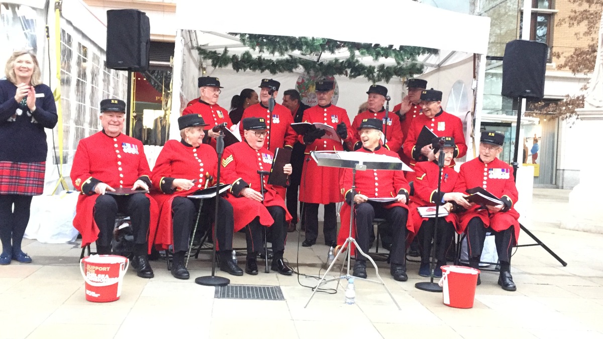 A choir of Chelsea Pensioners, a mixture of men and women who are smiling while wearing red jackets with large round buttons and many medals, along with black trousers. Their female conductor, smiling and applauding them to one side, is wearing a Christmas jumper, a tartan skirt and black tights.