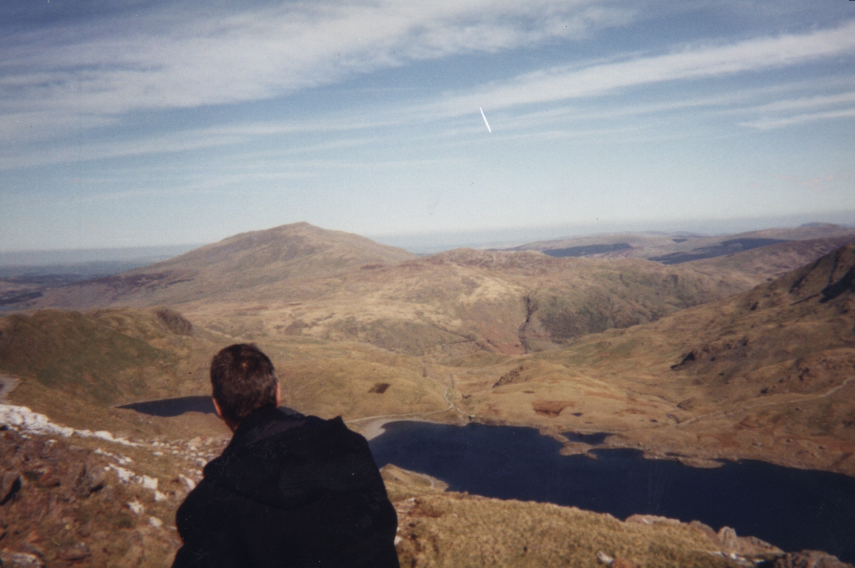Standing on a hill high above a lake, a man looks out over the beautiful mountainous scenery of Snowdonia, under a blue sky with wispy clouds.