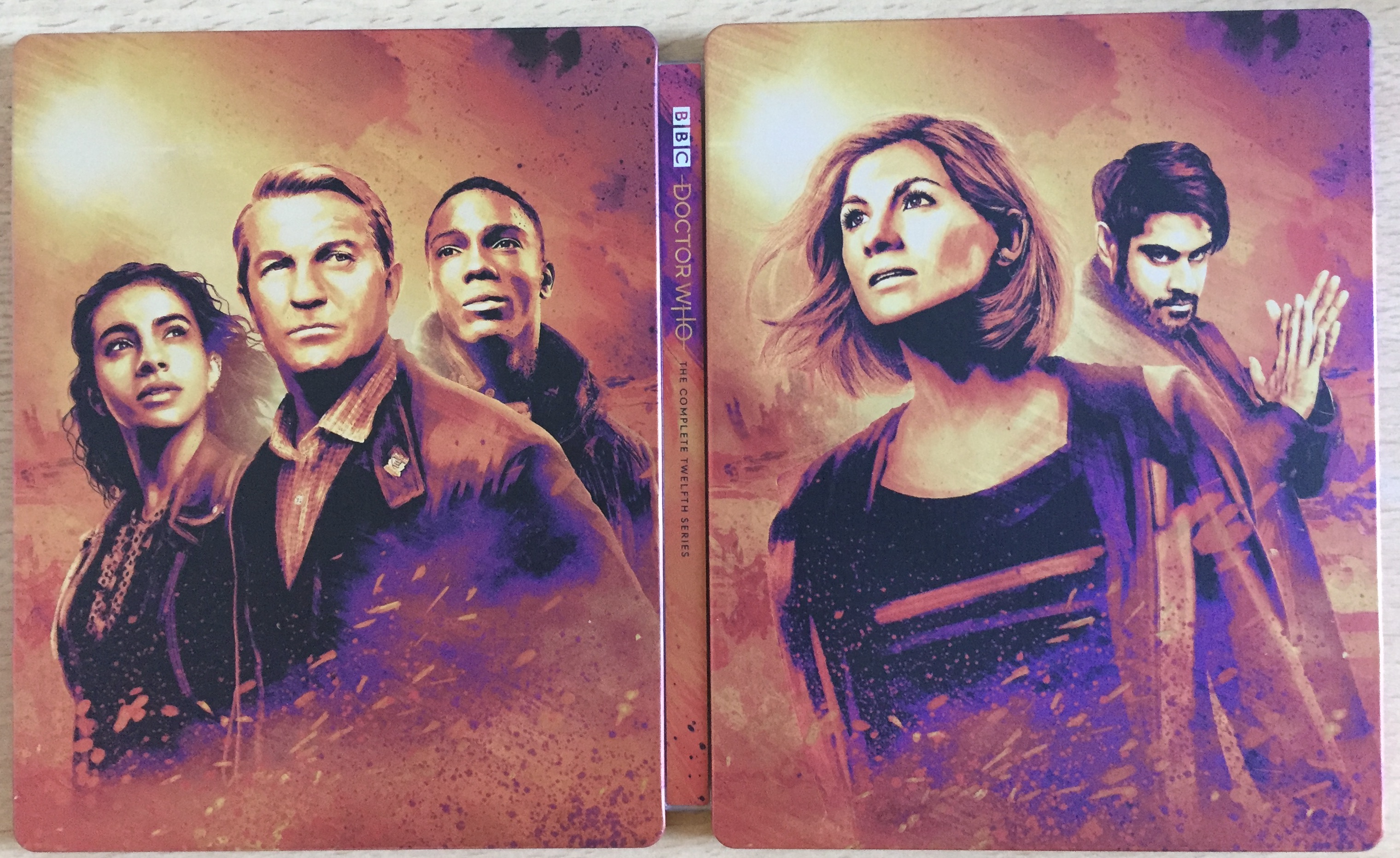 Steelbook cover for the blu-ray set of Doctor Who Series 12, spread open so that the artistic portraits of the cast members on the back and front covers are both visible. On the right, the front cover shows the Doctor and the Master, while the back cover on the left shows companions Yasmin, Graham and Ryan.