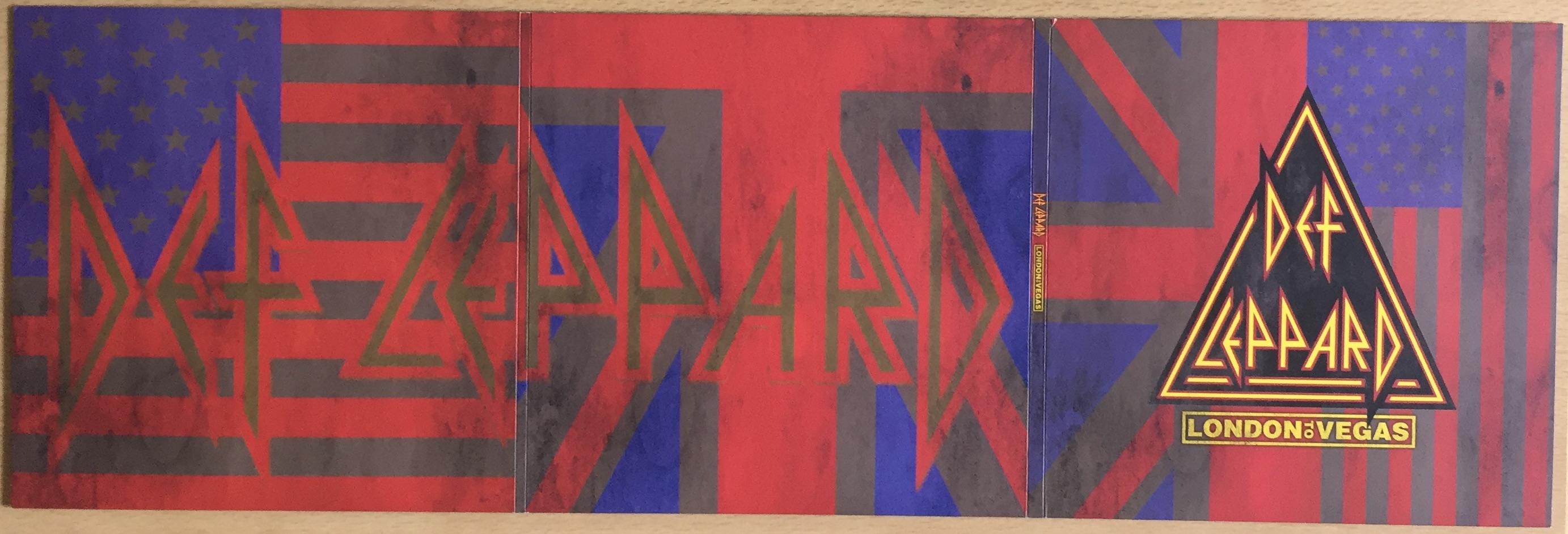 Outer side of the large 3 panel foldout sleeve from the Def Leppard London To Vegas box set. It features faded versions of the Union Jack and American flags across the background, the white areas having become grey while the reds and blues are still visible. A bit of dirt has been added to the flags as well. Across the left 2 panels, the Def Leppard logo spreads all the way across in large faded letters that are difficult to see. The far right panel, which forms the front of the sleeve when folded, as the Def Leppard logo in its more traditional clear form, inside the yellow and red edged triangle, with London To Vegas written beneath.