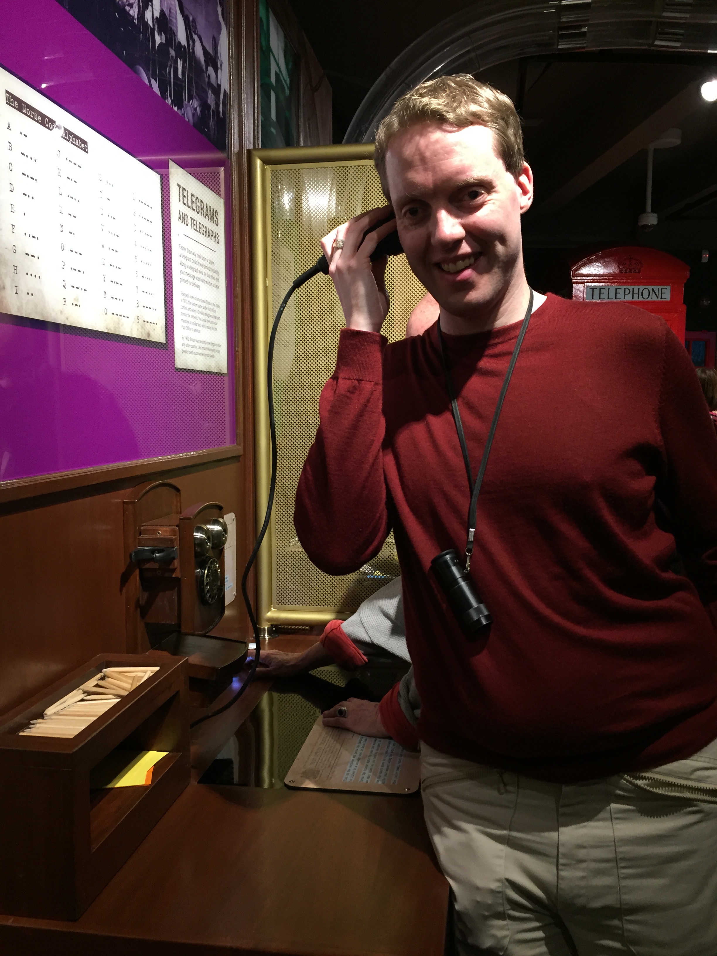 Glen smiling as he holds the receiver of a morse code device to his ear in a museum.