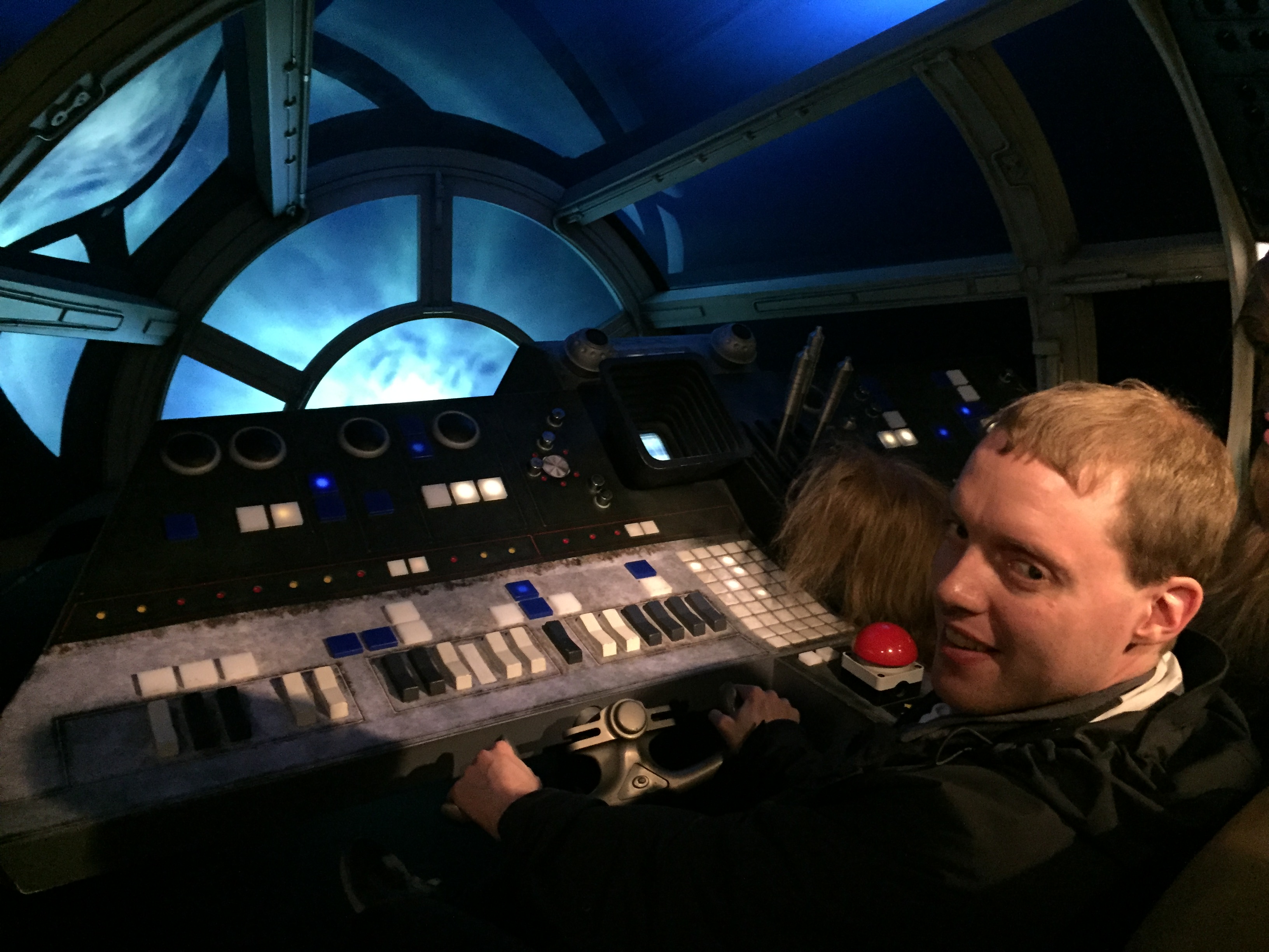 Glen sitting and grasping the steering control in the cockpit of the Millennium Falcon, the dashboard of which has black, white and blue chunky, tactile buttons that contrast well against the grey surface. Light outside the cockpit window gives the impression that he's zooming through space.