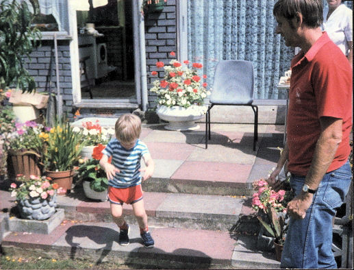 Glen as a child walking down a few paved steps from a patio into a garden, while his Dad watches.