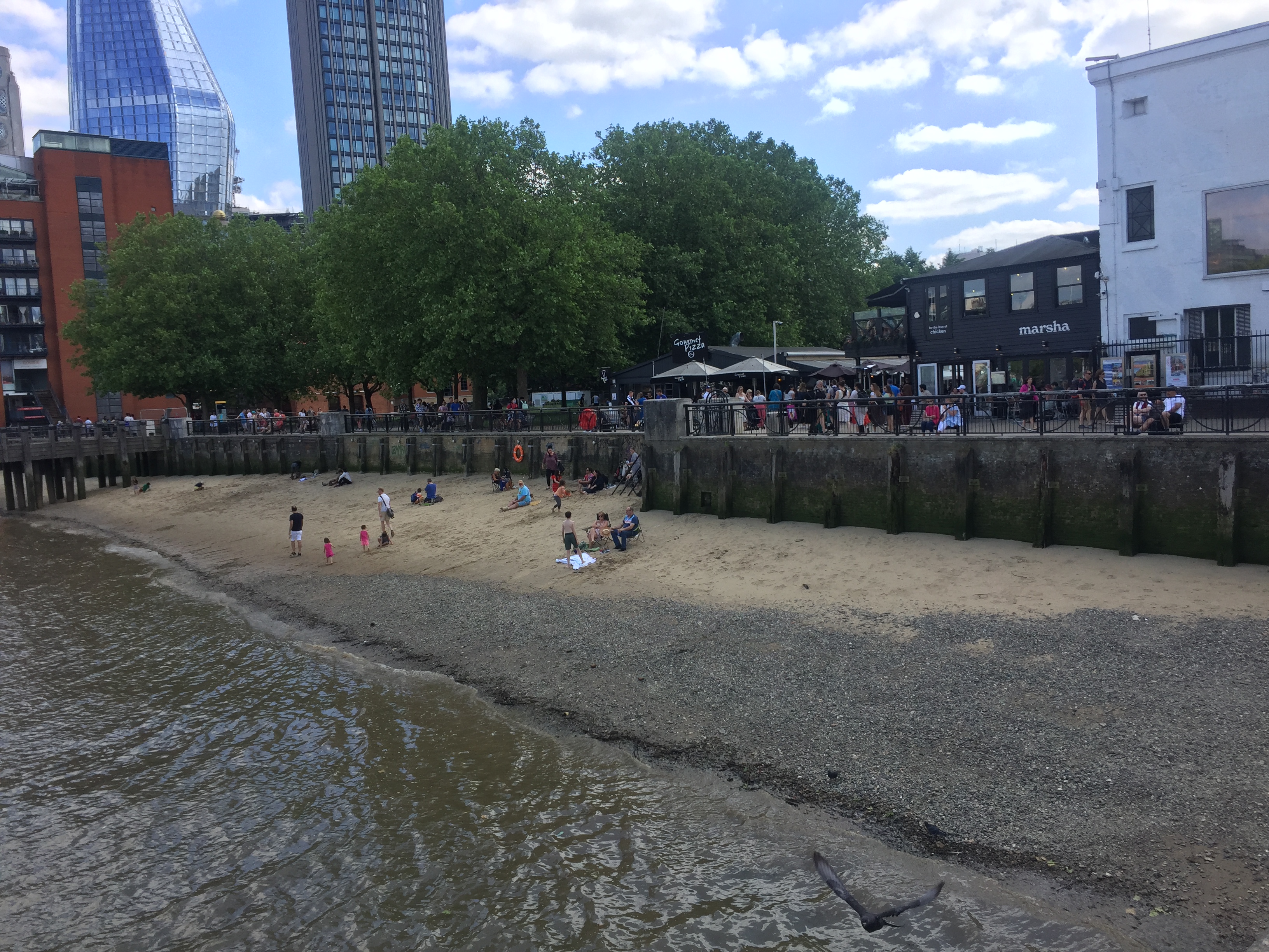 Adults and children on a small beach by the Thames, while lots of people walk along the main path behind them.