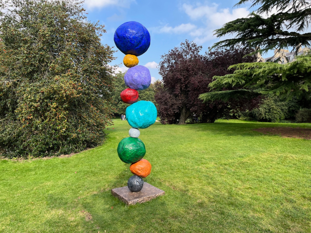 Tall sculpture made of different coloured boulders, unevenly stacked on top of each other in alternating small and large sizes. Colours include blue, green, organge and red. The sculpture is standing on a square panel on the grass, with trees in the background and wispy clouds in the blue sky above.