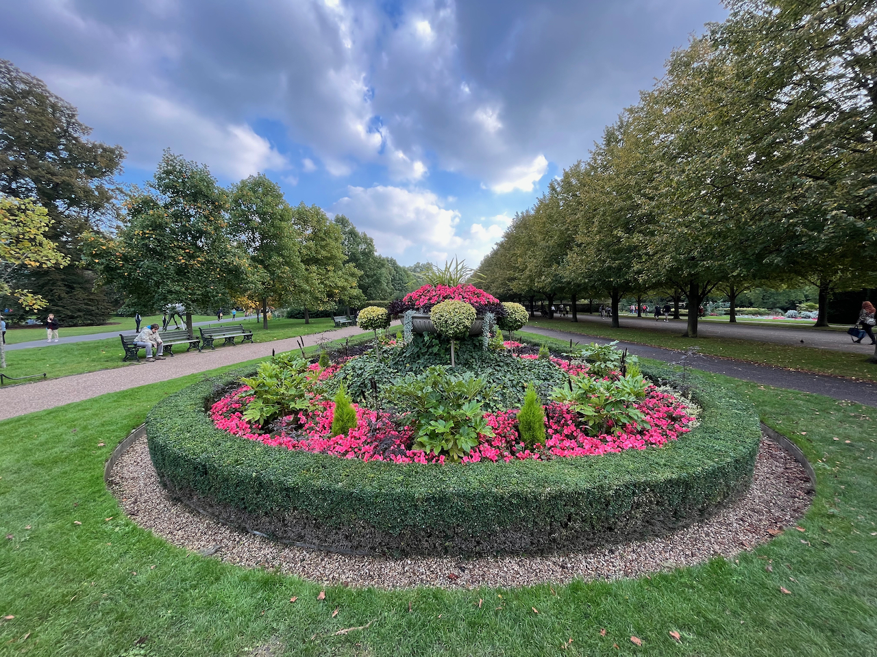 Large flowerbed stretching into the distance, surrounded by a low green border hedge that curves round at the end. The display contains red flowers and assorted greenery, including small trees with thin stems and round leafy heads, plus a large round metal planter containing more red flowers. Trees and benches line the paths on each side of the flowerbed, while above it all the sky is a dramatic mix of grey and white clouds with patches of blue sky.