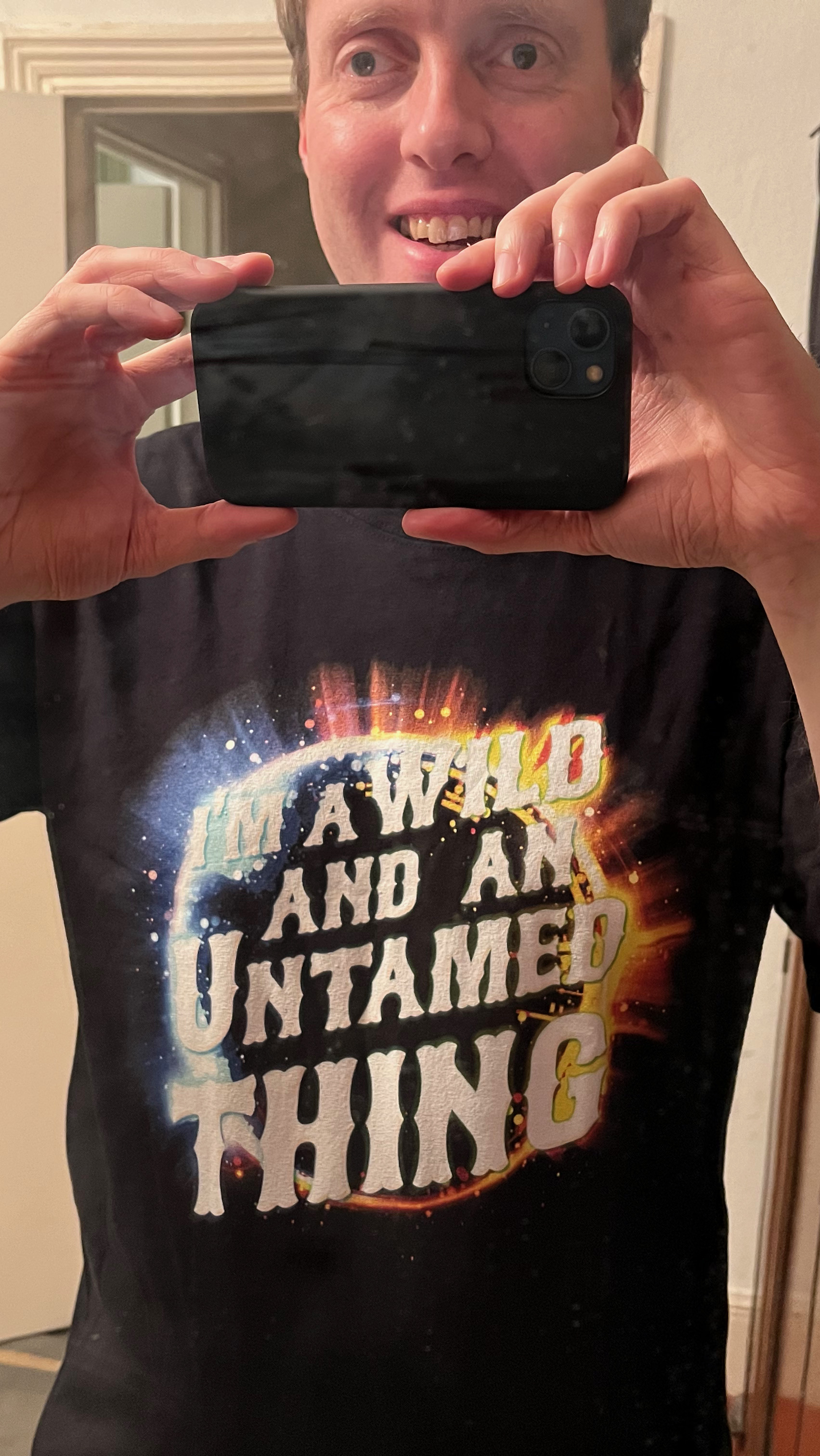 A mirror selfie of me wearing a black t-shirt that says I'm A Wild And Untamed Thing, in big white letters. Behind the text is a circle with a multi-coloured border that is radiating out lines and spots, as if giving off energy or exploding.