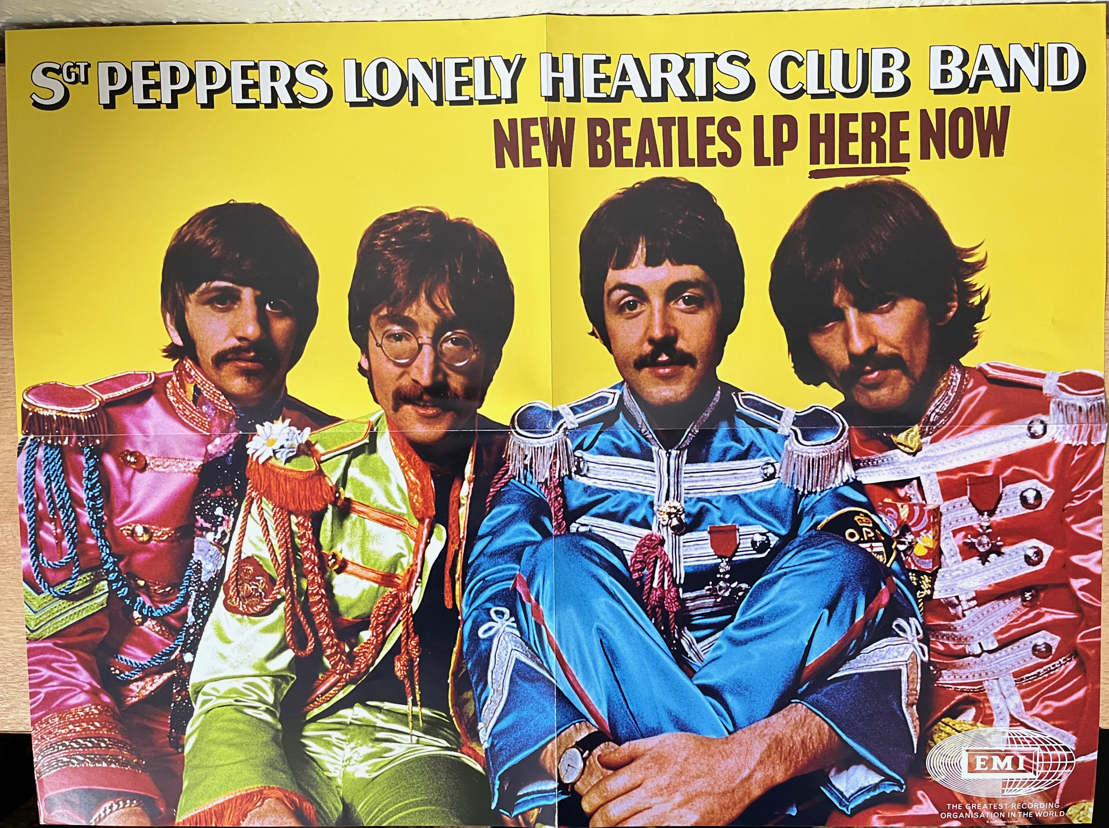 Poster of the 4 members of The Beatles, dressed in their ornately decorated pink, yellow, blue and red Sergeant Pepper outfits, against a yellow background, below the text Sergeant Pepper's Lonely Hearts Club Band, New Beatles LP here now.