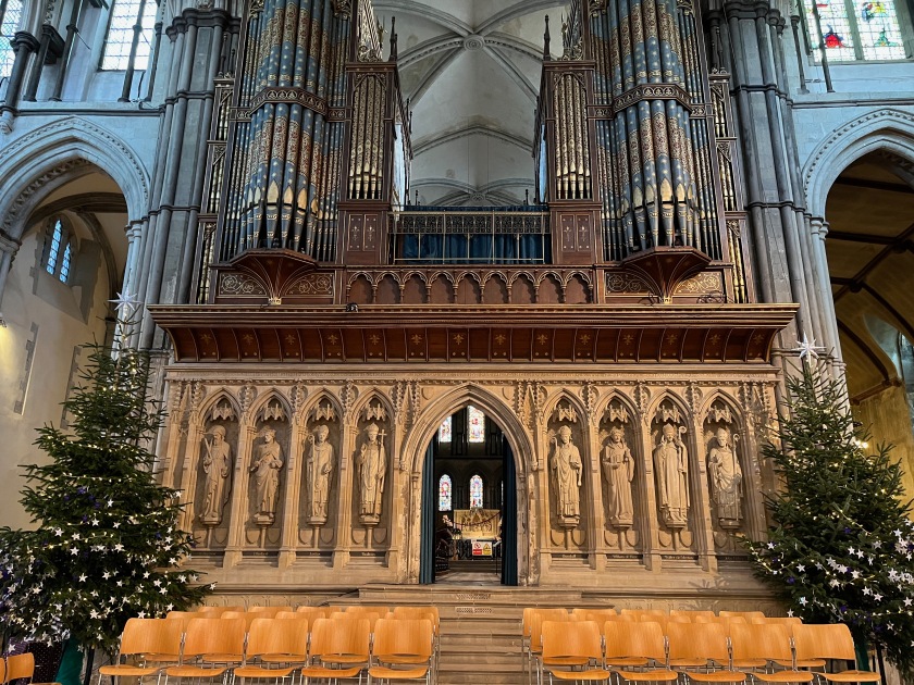 Below the huge ornate pipes of the cathedral organ, 8 statues embedded in narrow arches stand in a line, separated halfway along by a larger archway into another room. Decorated Christmas trees stand on the far left and right of the row of statues.