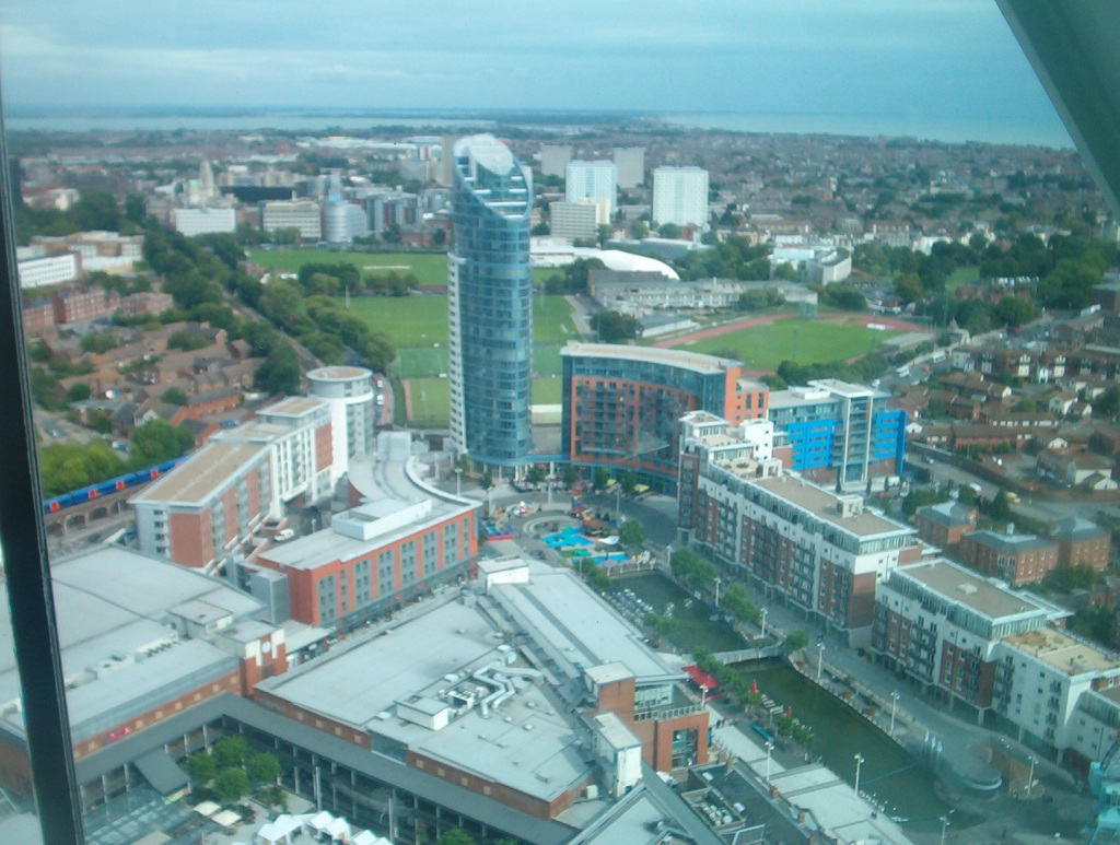 View from the Spinnaker Tower towards a high skyscraper with a steeply sloping roof, surrounded at ground level by apartment blocks, and a large green park behind it.