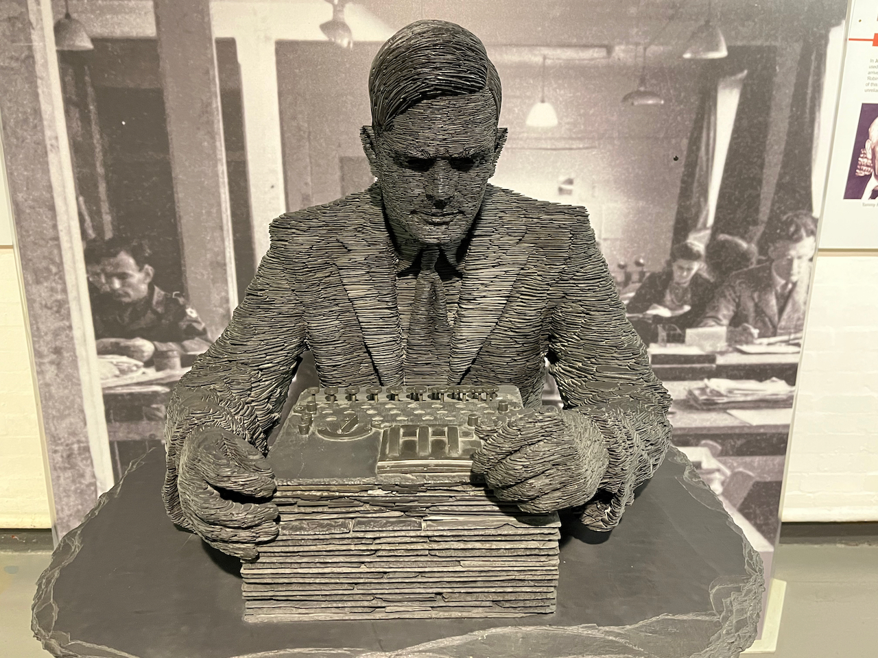 A closer view of the Alan Turing statue, showing his head and upper body, and the Enigma machine he's holding on the desk.