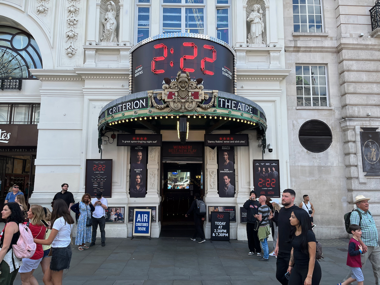 The exterior of the Criterion Theatre. The curved canopy overhanging the entrance has an ornate grinning figurehead centred in between the words Criterion and Theatre. Above that, in large red numbers against black, is a digital clock face showing 2:22. Either side of the entrance doorway below, meanwhile, are vertical boards with the headshots and names of the 4 lead actors against a black background, 2 on each side.