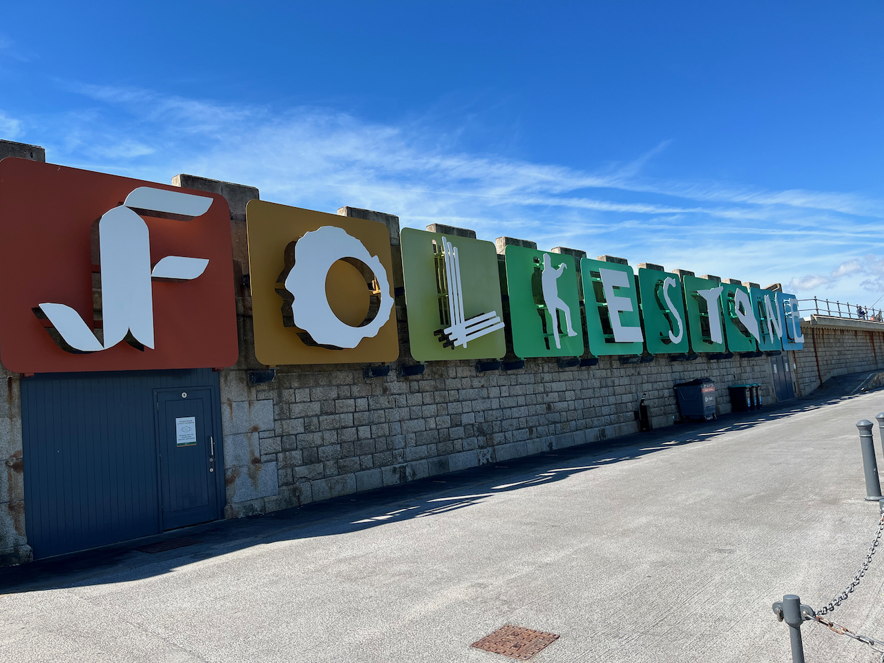 A long wall sign made up of very large coloured blocks that spell out the word Folkestone in stylised white letters. Each letter is styled differently, with the K in particular looking like a person holding up their left arm and leg to form the letter.