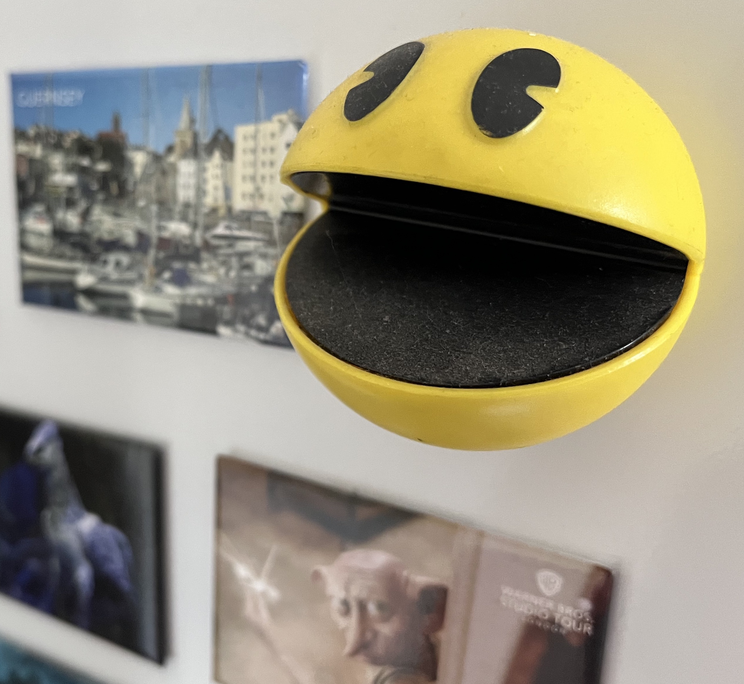 A fridge magnet in the shape of Pac-man. The magnet is a yellow ball that sticks out beyond the flat magnets next to it. It has a large black wide open mouth across its width, and a couple of black coloured eyes just above it.