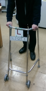 A metal zimmer frame with 2 small pieces of paper attached to the handlebar at the top, a square piece with a letter L on it, and a rectangular piece below it, like a registration plate, with the text Old Git 1.