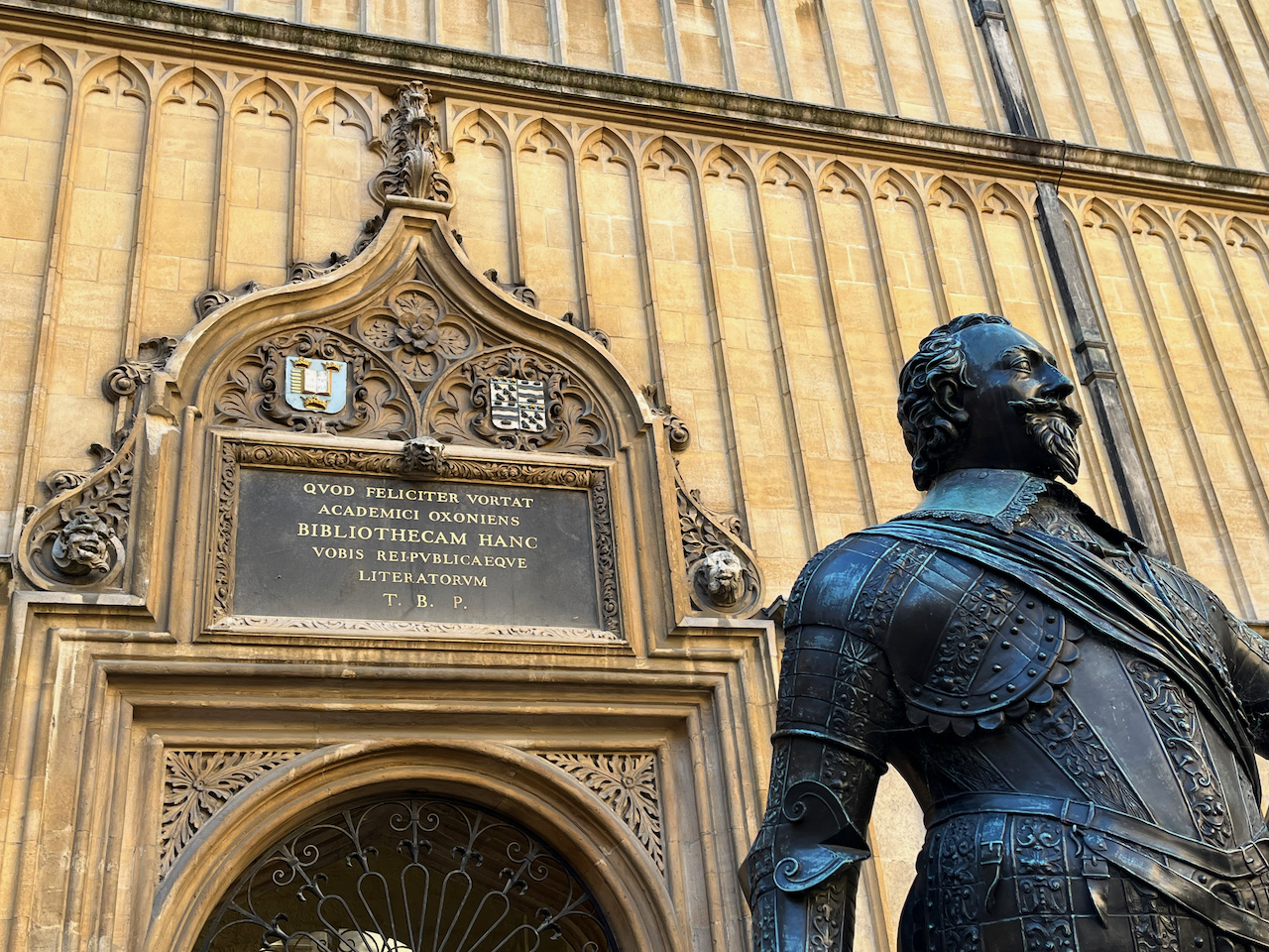 Latin text, decorative shields and ornate carving above the entrance to the Divinity School, with the statue of the Earl of Pembroke in front of it.