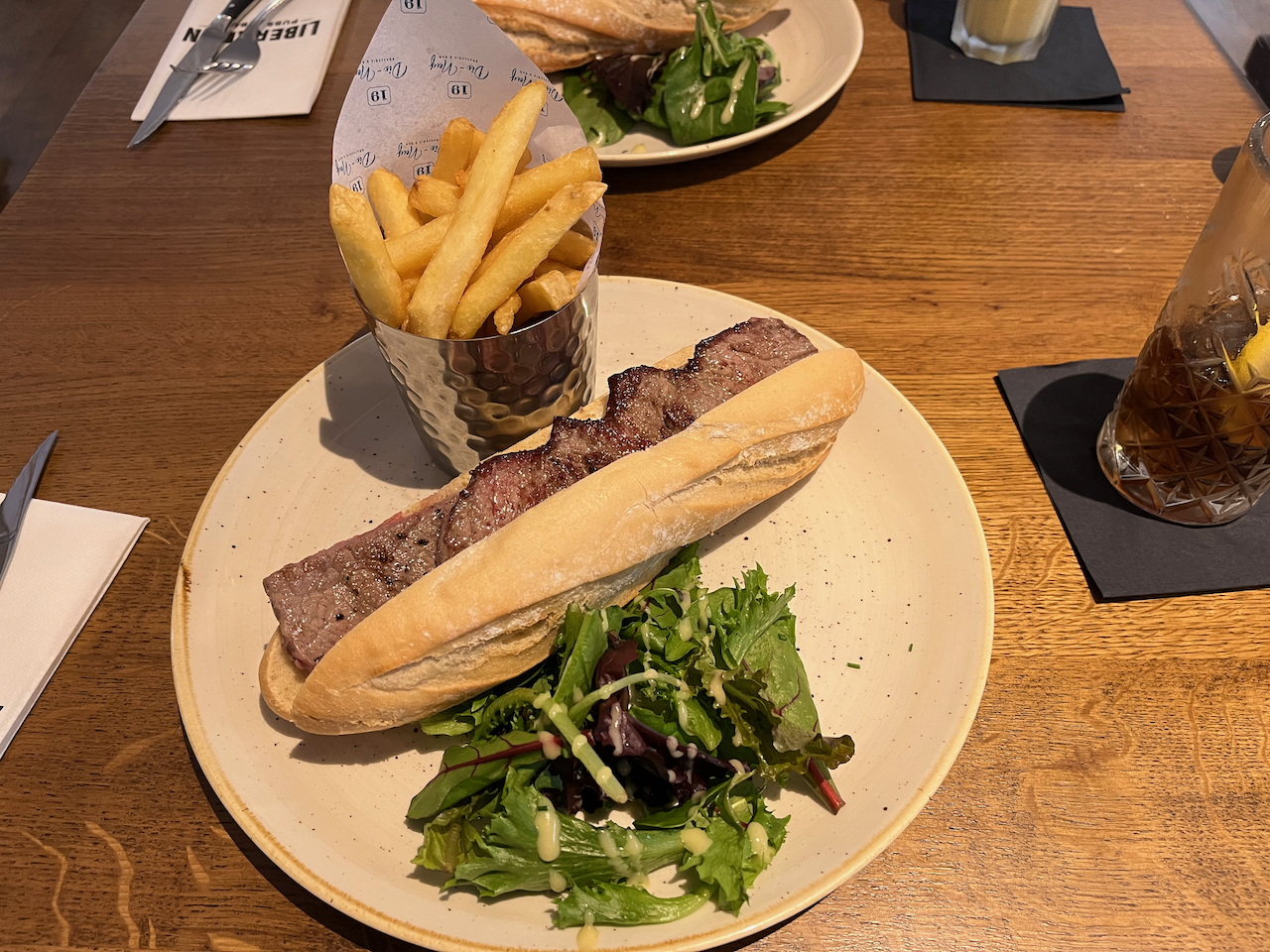 A large steak baguette with green salad leaves on a plate, along with a metal can containing lots of large chips.