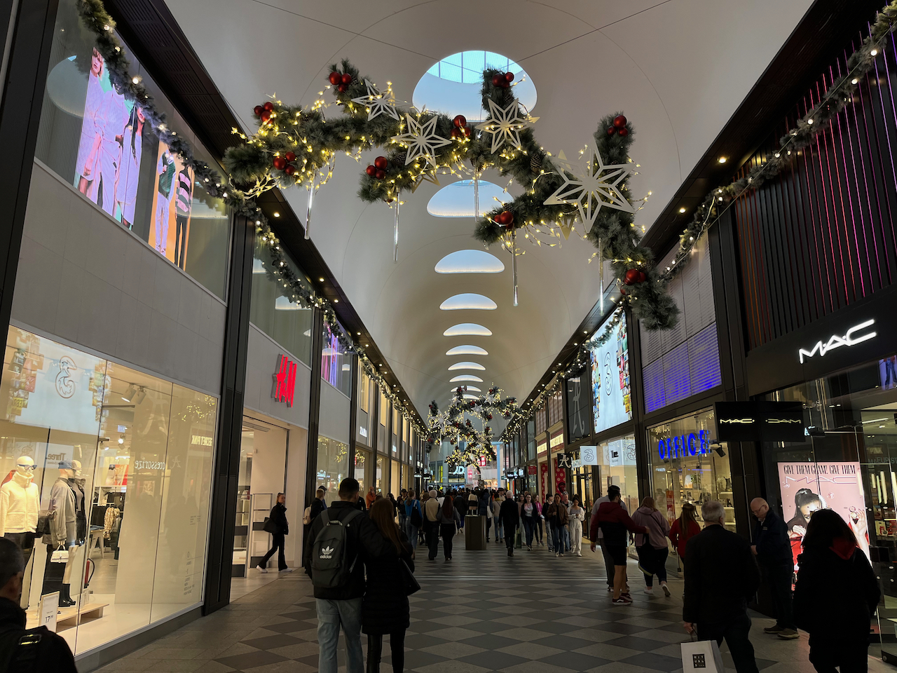 Christmas decorations hanging from the ceiling in a corridor of Westgate shopping centre, consisting of large branch-like shapes covered in greenery and lights, while more lights and tinsel are strung high along each wall of the corridor above the shop entrances.