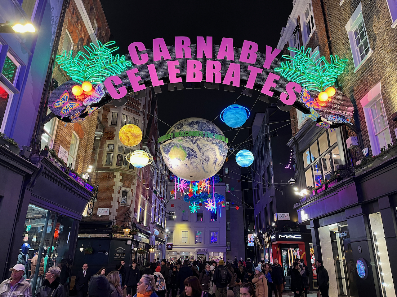 An archway with the words Carnaby Celebrates in big pink letters, with mistletoe-shaped Christmas lights and butterflies at each end. Beyond it, a large globe of planet Earth is suspended in the air with smaller planets orbiting it. Green text around the Earth reads Carnaby welcomes the world.