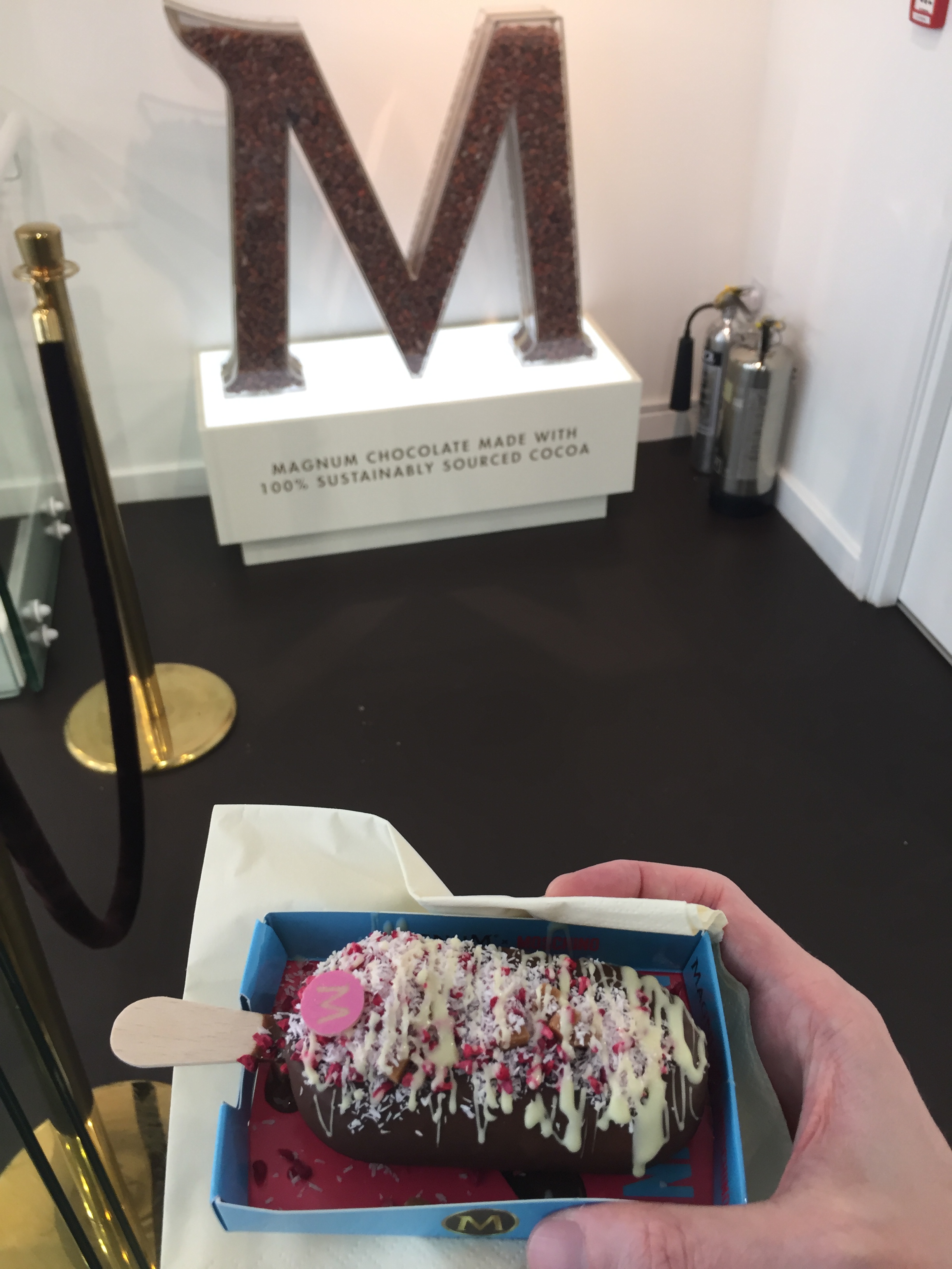 My custom designed Magnum, with double layered milk chocolate, coconut shavings, raspberry pieces, caramel fudge, raspberry white chocolate coins, and white chocolate drizzle. It's being held in front of a sign in the shape of a large letter M, under which text says Magnum chocolate made with 100% sustainably sourced cocoa.