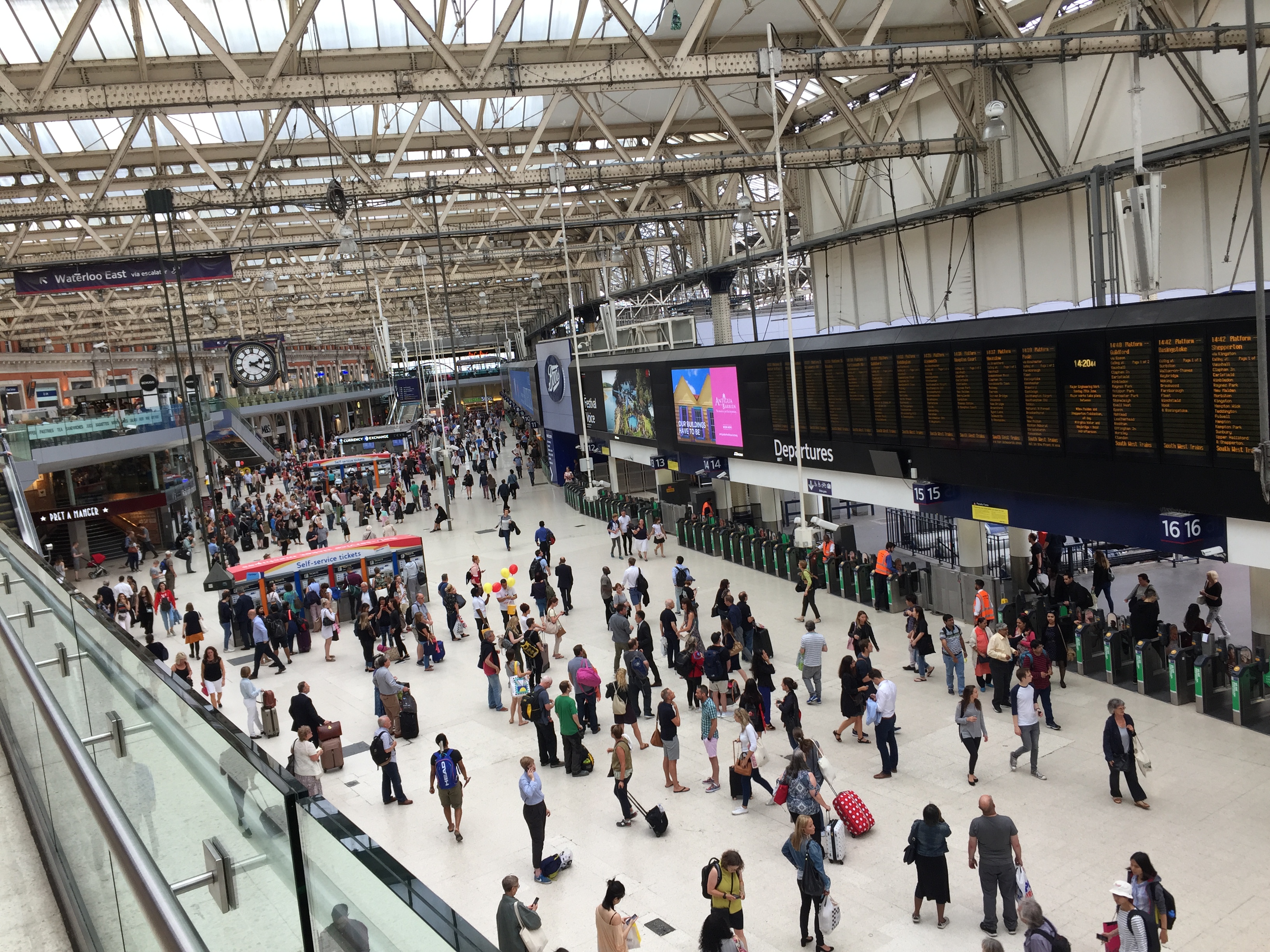 A wide few of the very busy concourse at Waterloo Station, as viewed looked down from the upper level, with ticket machines, ticket barriers, departure boards, advertising screens, and the iconic clock that hangs from the ceiling.