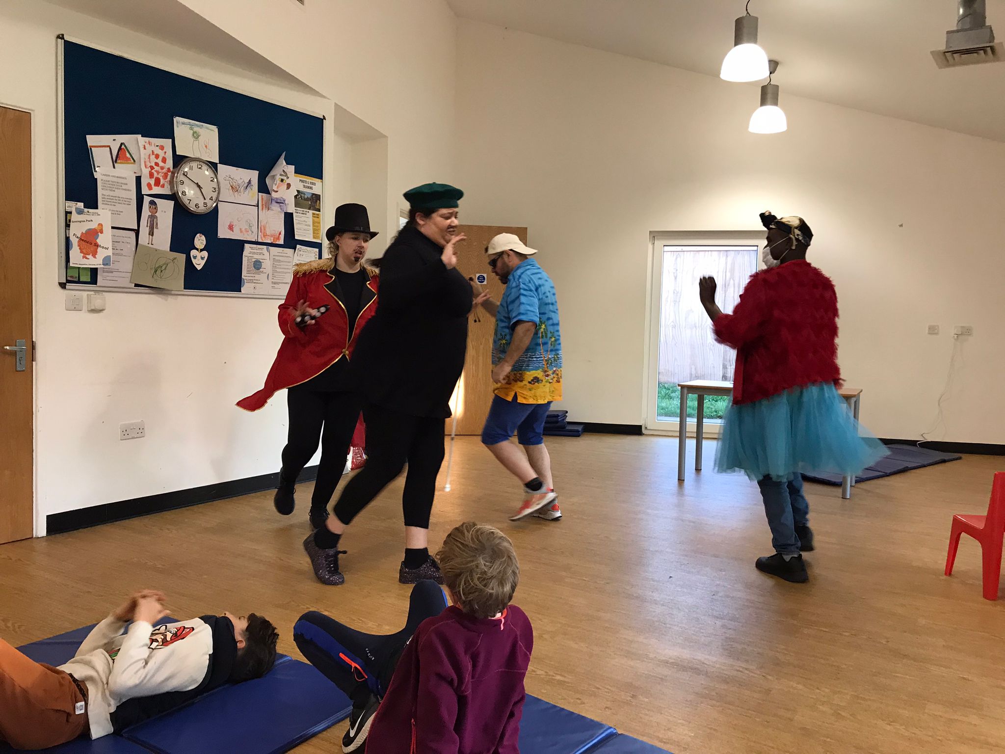 4 characters from Super Power Panto running around in a circle while 2 children watch them.