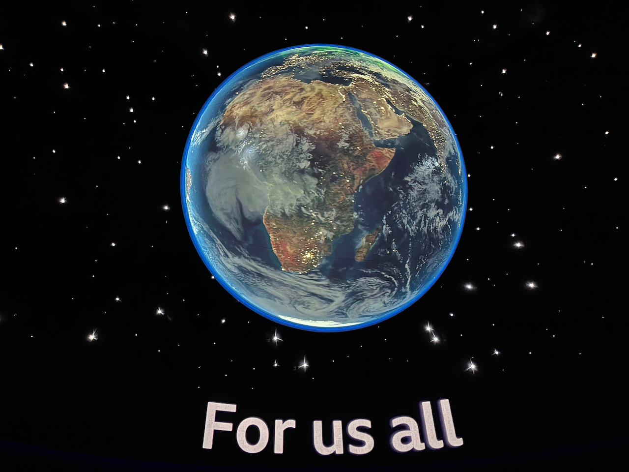 An image of planet Earth in space, surrounded by stars, above the words For us all.