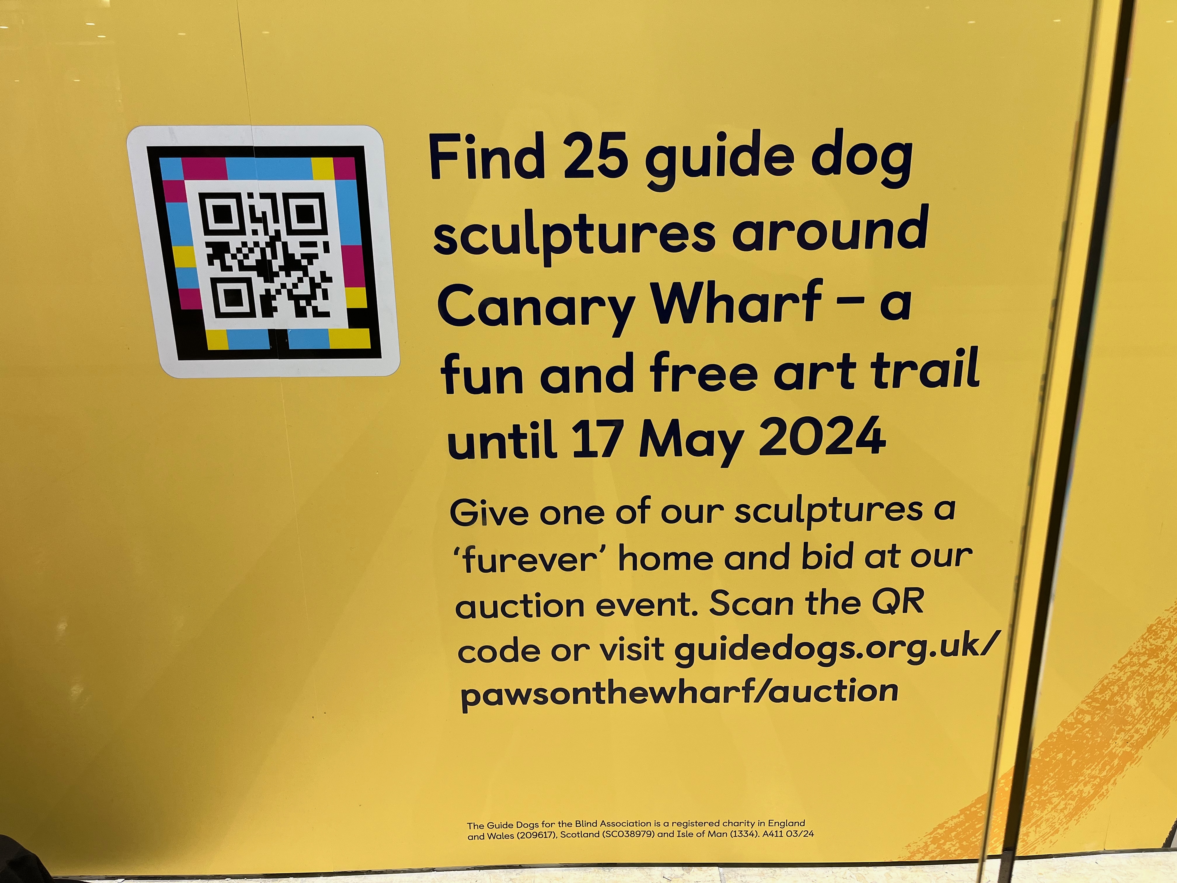 Against a yellow background, next to a black and white QR code with colour around its edge, is black text that says Find 25 guide dog sculptures around Canary Wharf - a fun and free art trail until 17 May 2024. Give one of our sculptures a 'furever' home and bid at our auction event. Scan the QR code or visit GuideDogs.org.uk/PawsOnTheWharf/Auction